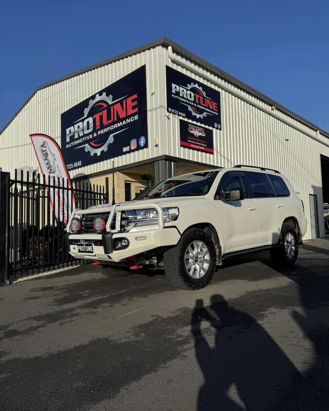 A white SUV parked outside a building with signs reading PRO TUNE AUTOMOTIVE & PERFORMANCE. The vehicle has off-road tires, a bull bar with lights, and a winch. The building has a black fence, banners, and signs for the automotive shop, under a clear