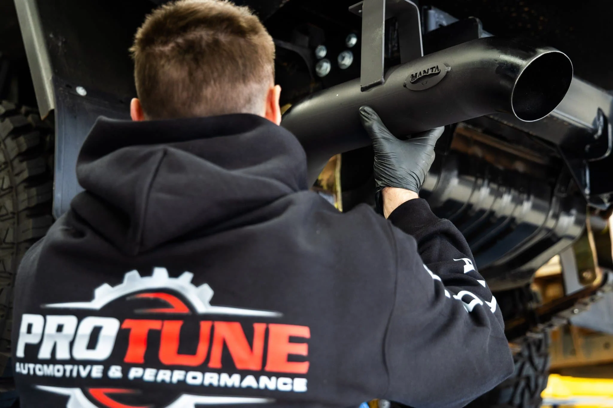 A person wearing a black hoodie with 'ProTune Automotive & Performance' logo, working on the exhaust system of a vehicle, holding a muffler in a garage or workshop.