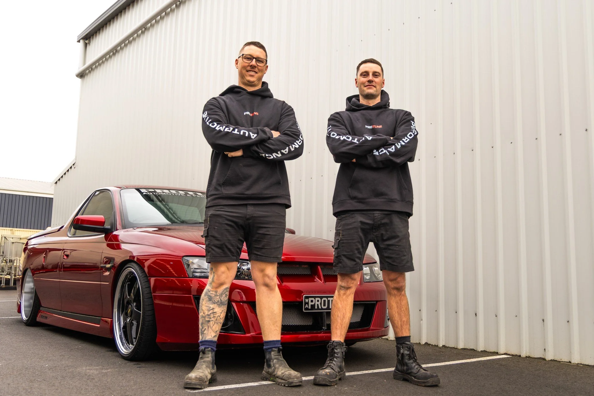 Two men in black hoodie sweatshirts and shorts standing with crossed arms in front of a red modified car with black wheels, parked outside near a beige metal building.