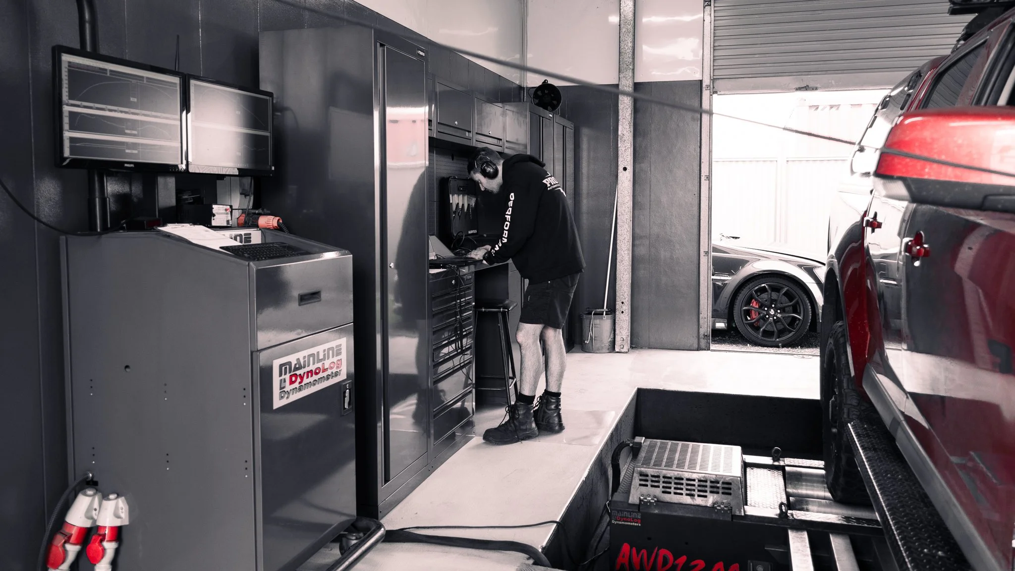 A mechanic working in a garage, surrounded by cars and diagnostic equipment, with two computer monitors and a technician's station.