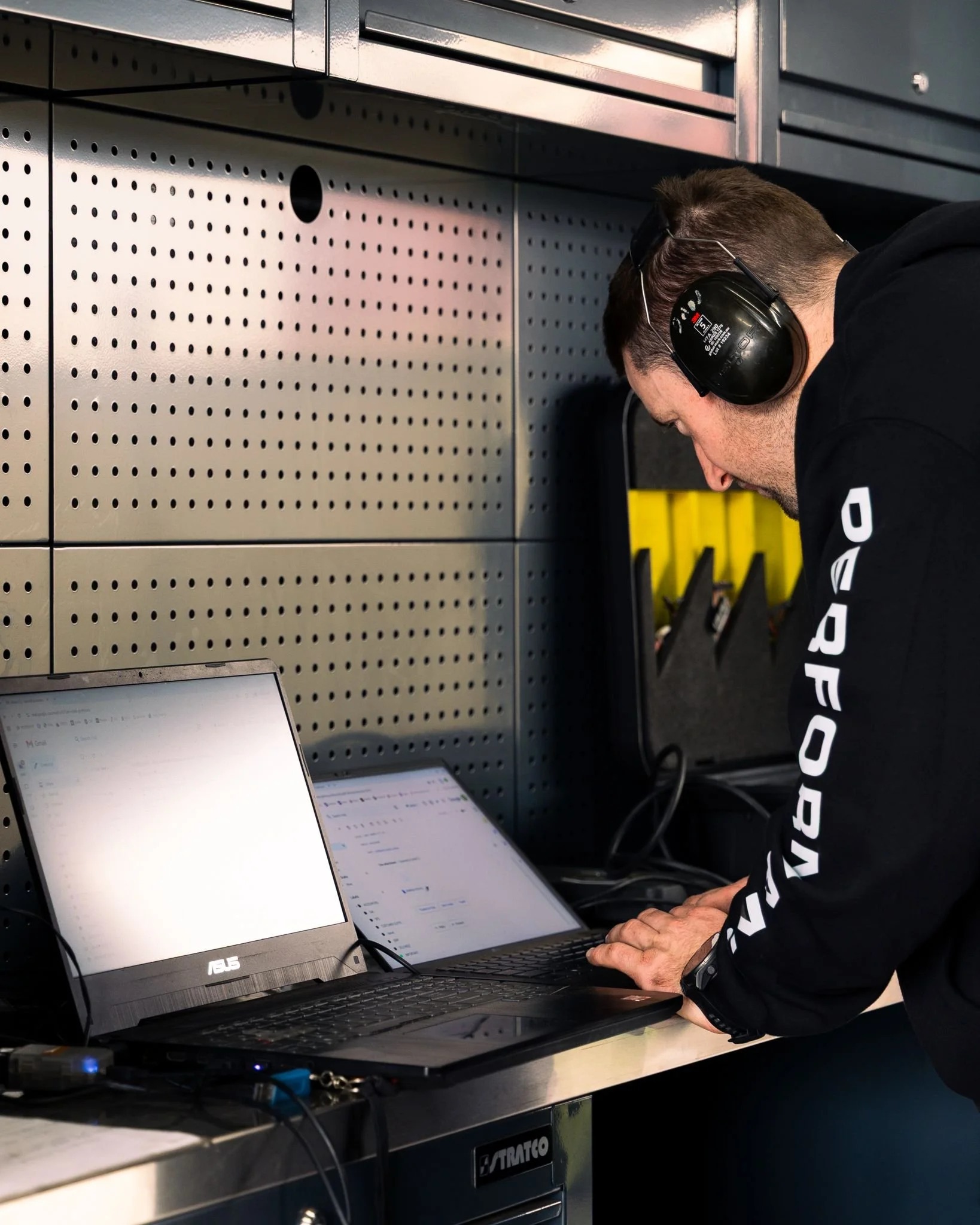 A man with headphones working on a laptop in a workshop or industrial setting.