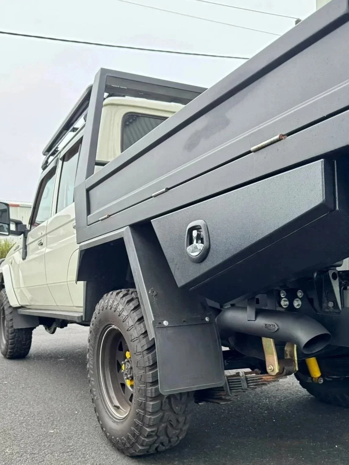 A close-up view of the rear side of a white 79 land cruiser with a black flatbed, including its large off-road tire, mud flap, and part of the black exhaust system.