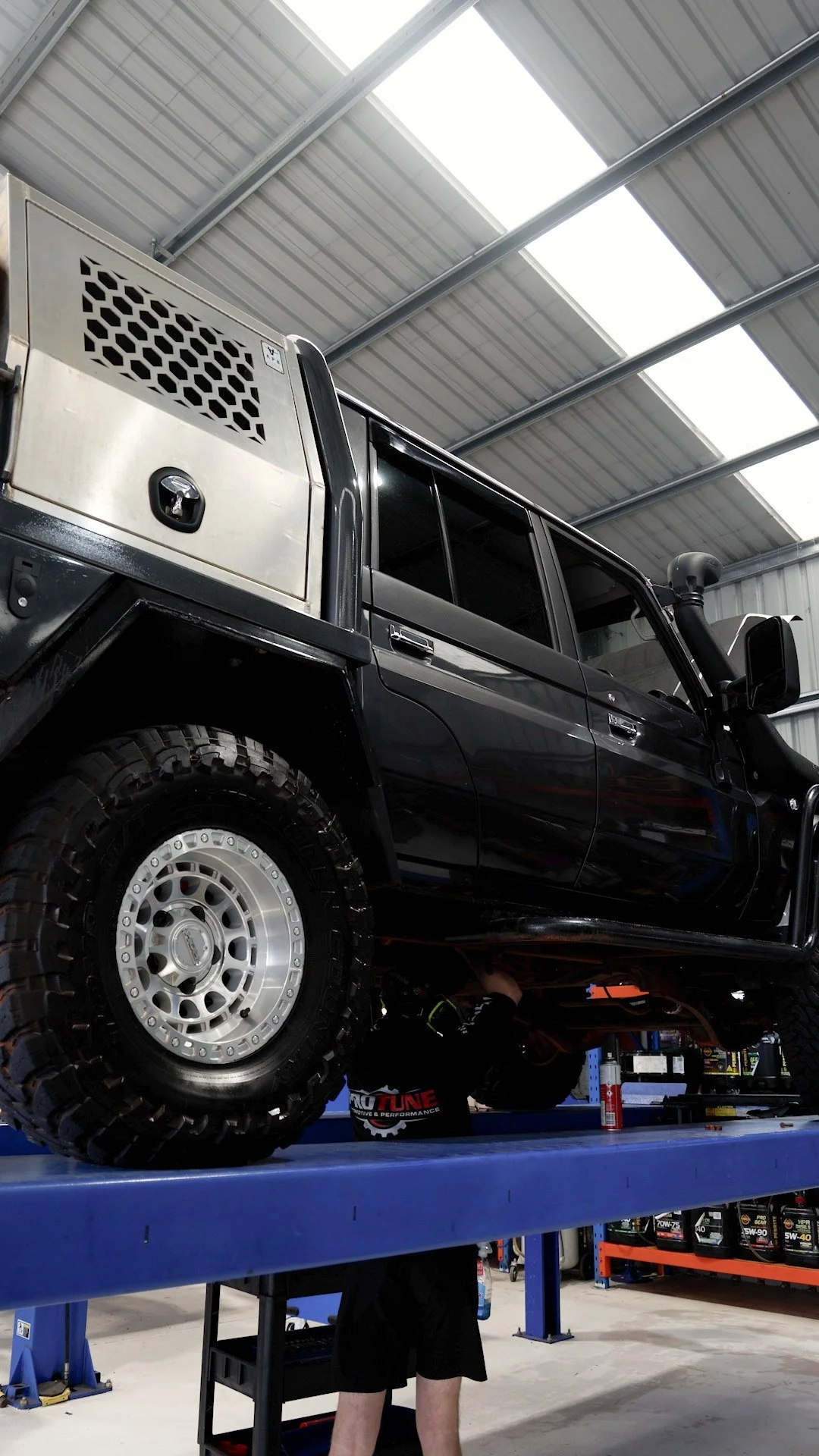 A black off-road vehicle is lifted on a blue service lift inside an auto repair shop. A mechanic is working underneath the vehicle.