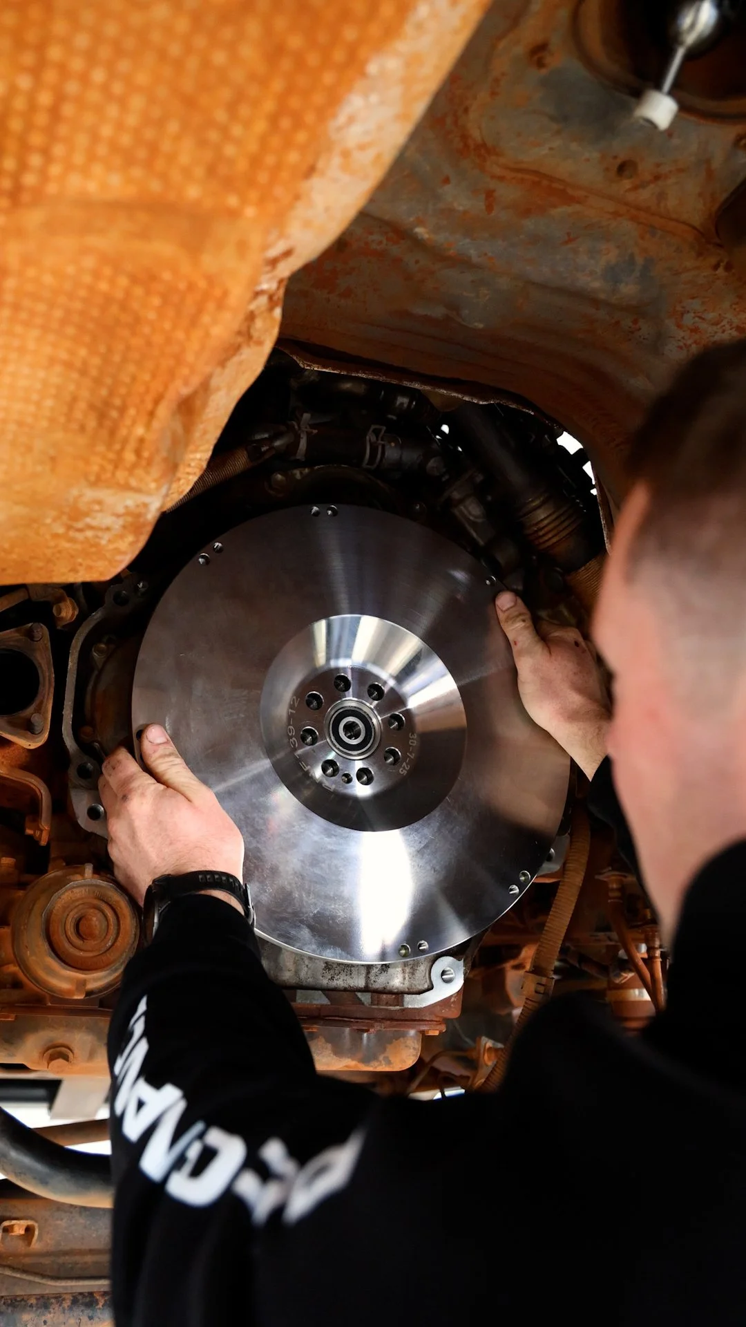 Person installing a shiny, circular metal flywheel inside a machine or engine compartment.