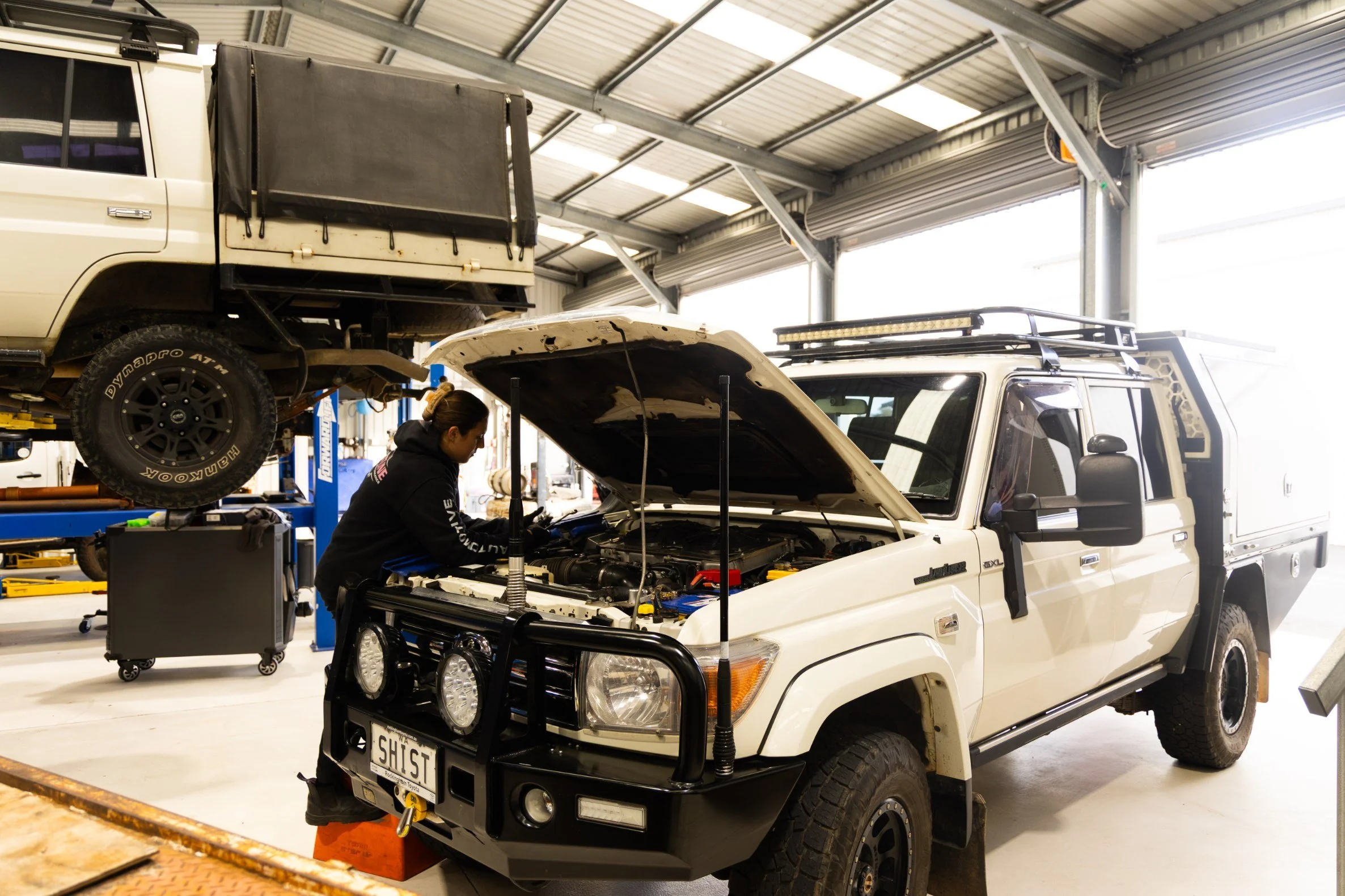 A mechanic working under the open hood of a white off-road vehicle inside a garage, with another vehicle lifted on a hydraulic lift in the background.