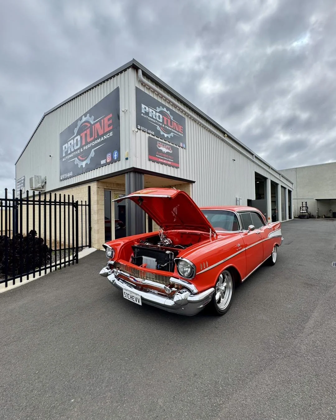 A red vintage Chevrolet car with open hood parked outside an automotive repair shop named PRO TUNE. The shop has a large sign with logos and contact info on the building. The sky is cloudy.