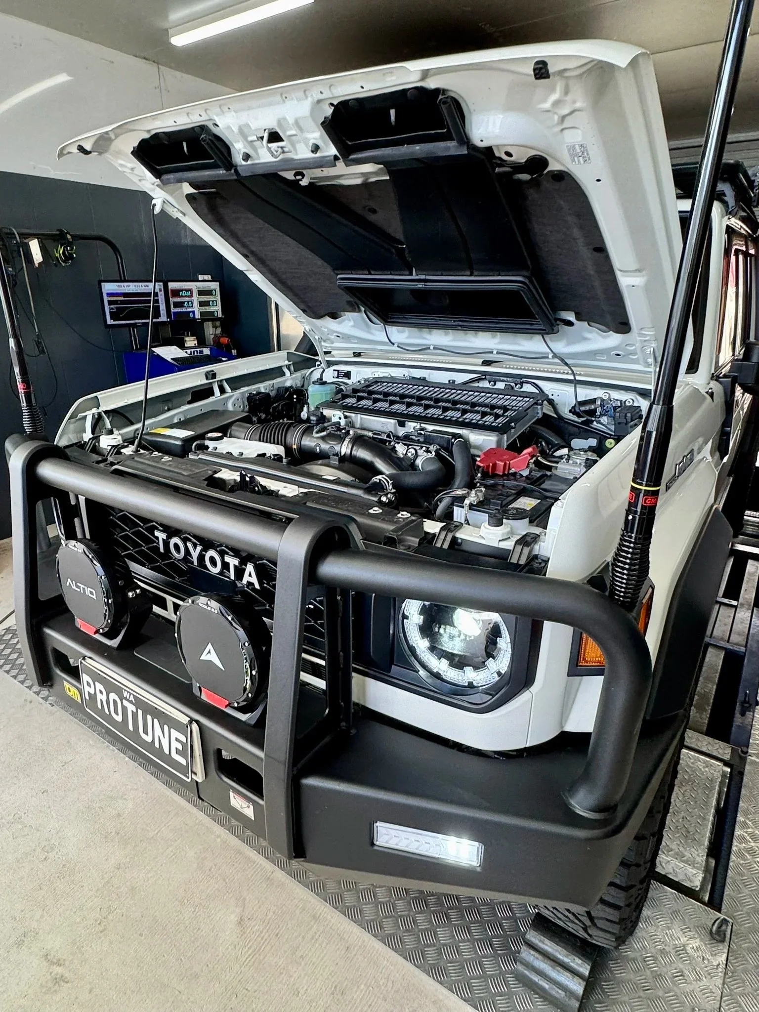 Under the open hood of a white Toyota vehicle, showing a clean engine bay with various engine components. Front bumper has a ProTune license plate and off-road lights, with a sturdy black grille guard and off-road shocks visible.
