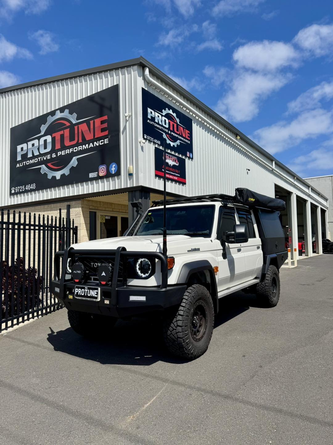 A white off-road vehicle parked outside of an automotive and performance shop named PRO TUNE. The shop building has a large sign with the company logo, contact information, and social media icons. The sky is partly cloudy.