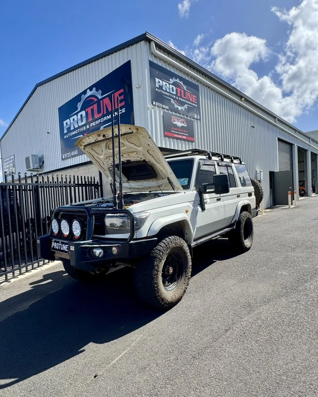 A white off-road vehicle with its hood open parked outside a building with signs that say PRO TUNE and Automotive & Performance, suggesting a car tuning or performance shop.
