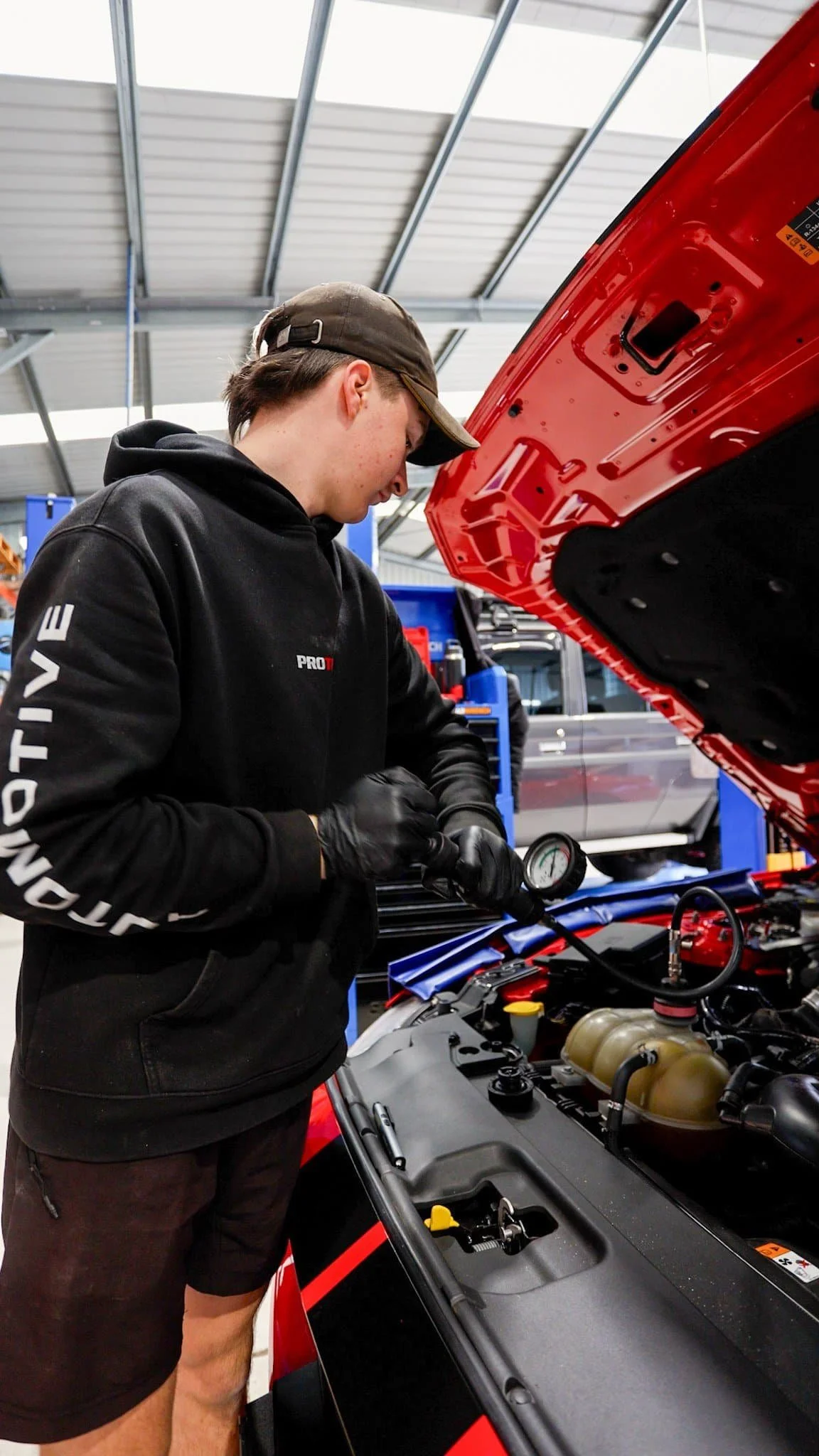 Two automotive technicians working on a vehicle with the hood open in an automotive repair shop. Both are wearing black hoodies with 'PRO TUNE' and 'Automotive & Performance' written on the back. Parts of a white off-road vehicle are visible in the background.