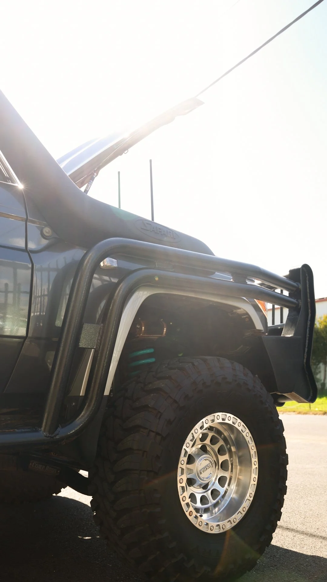 Close-up of the front left side of a black off-road vehicle with large tires, a bumper, and open hood, parked under bright sunlight.