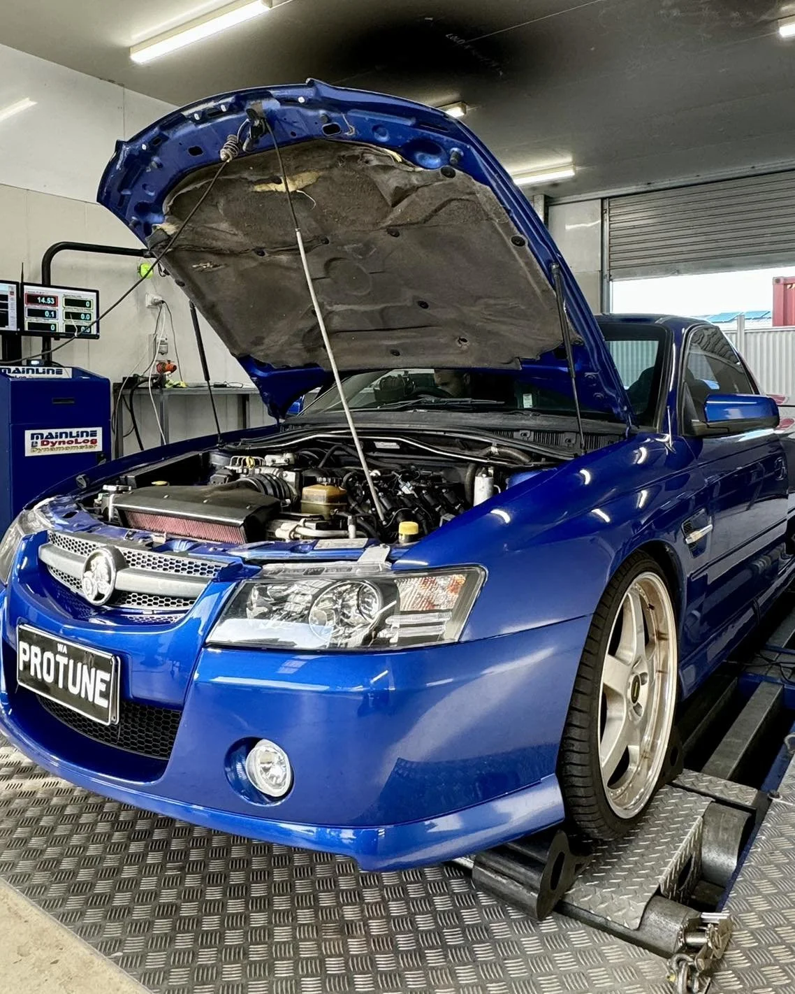 A blue car with its hood open on a dynamometer testing platform in a garage or workshop.