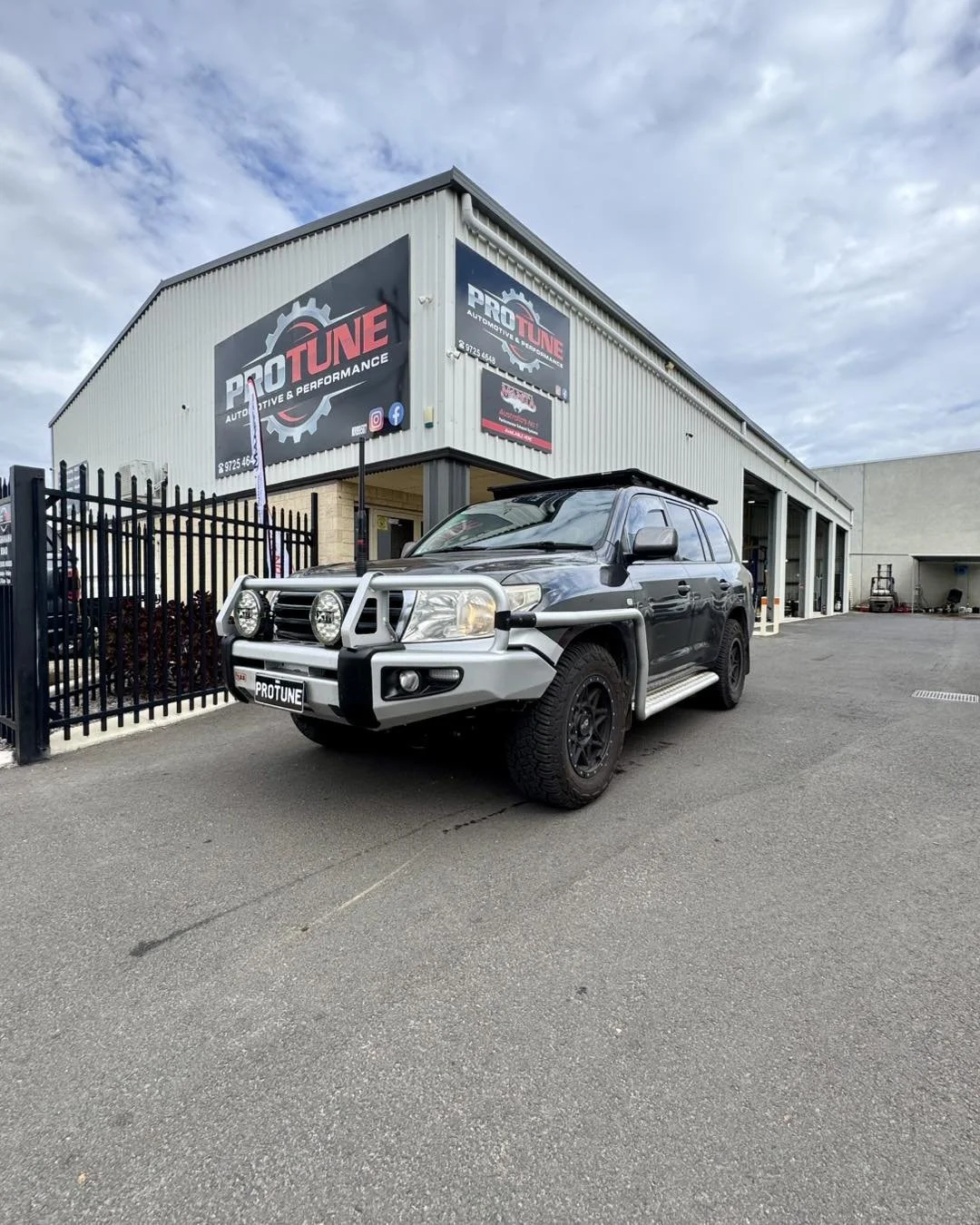 A black SUV with silver bull bars, two large off-road lights, and black wheels parked outside an auto performance shop called PRO TUNE, with a gray industrial building and cloudy sky in the background.