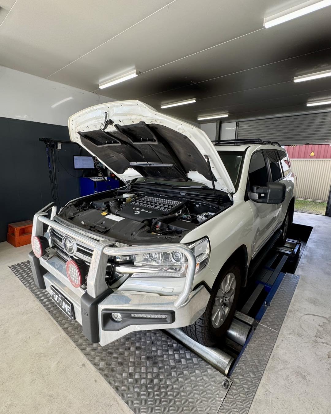 A white SUV with the hood open inside a garage or workshop, revealing the engine. The vehicle is on a roller testing platform, and the front is equipped with a bull bar with mounted lights. The garage has fluorescent lighting and some equipment in the background.