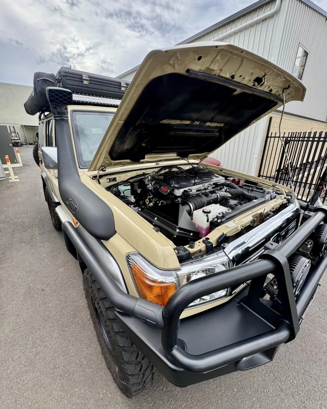 A beige 4WD vehicle with the hood open, showing the engine. The vehicle has large tires, a black front bumper guard, and a roof rack.