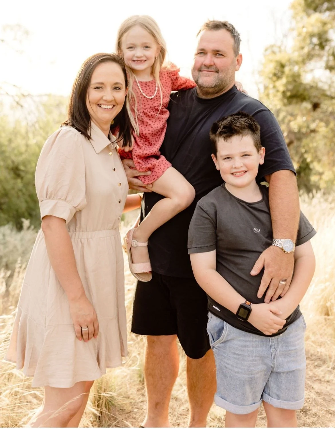 A family of four posing outdoors on a sunny day, with a woman, a man, a girl, and two boys, all smiling and standing on a grassy field with trees in the background.