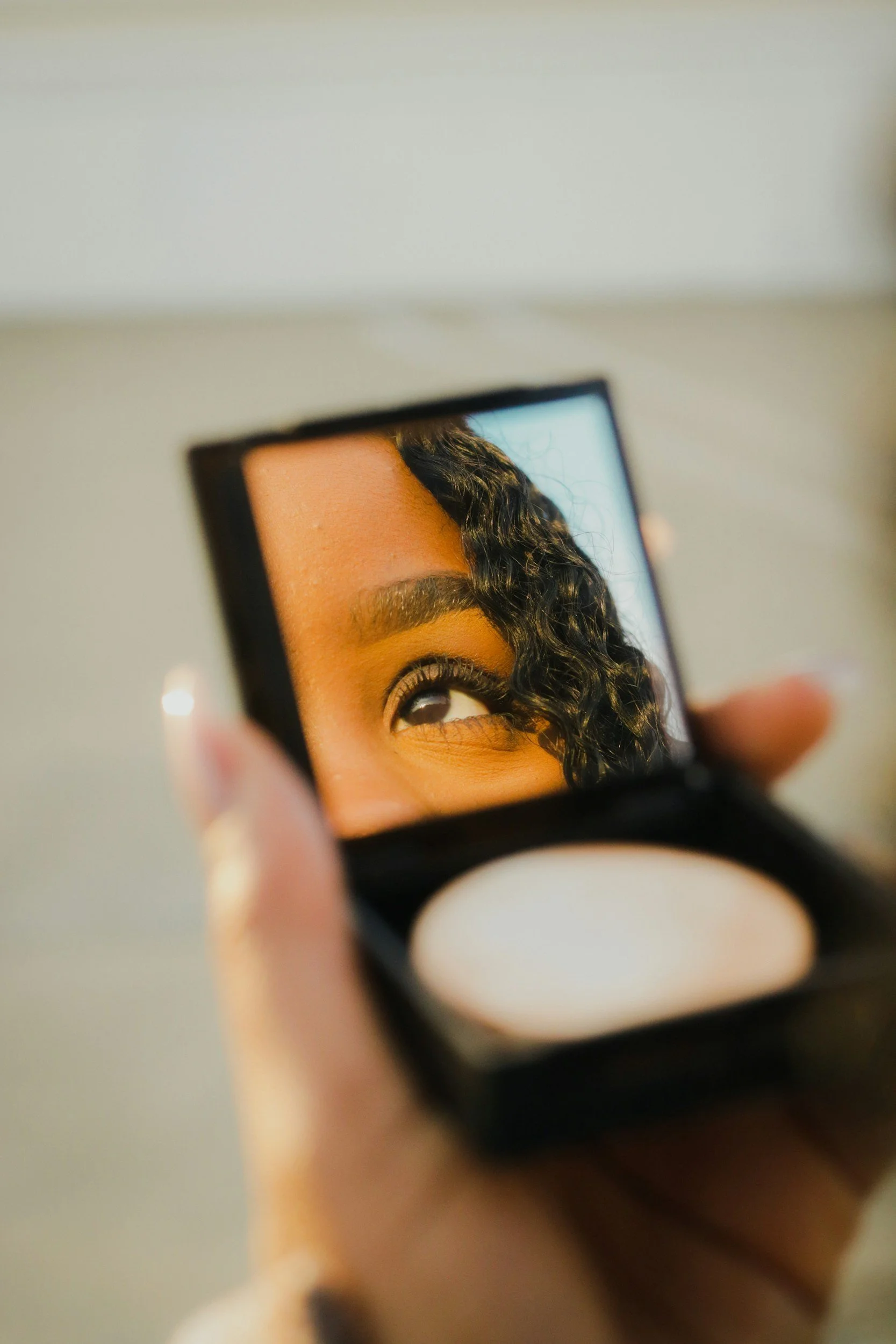 A person's eye and eyebrow reflected in a compact mirror, with curly hair partially covering the eye.