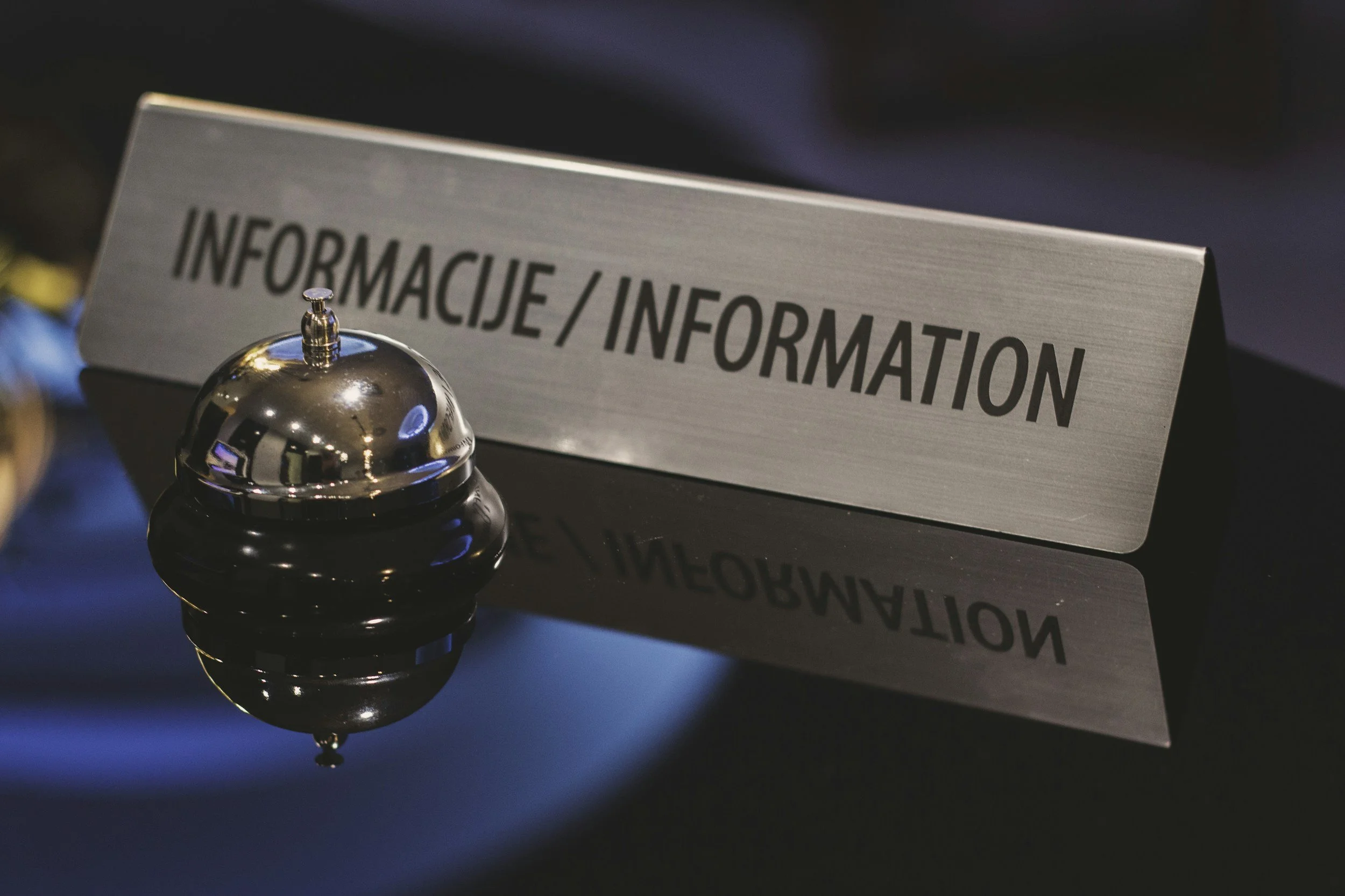 A metallic desk bell beside a silver sign with black text that reads 'INFORMACIJE / INFORMATION'.