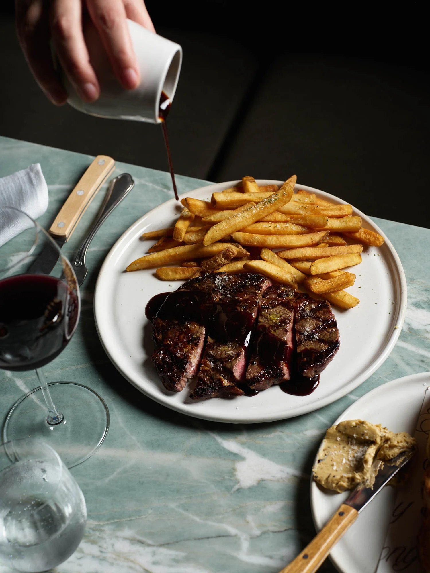 A plate of French fries and a sliced steak with dark sauce on a green marble table. Someone is pouring sauce from a small white cup onto the fries.