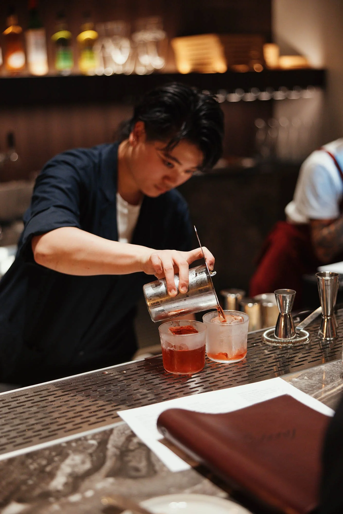 Bartender pouring a red drink into two glasses at a bar counter.
