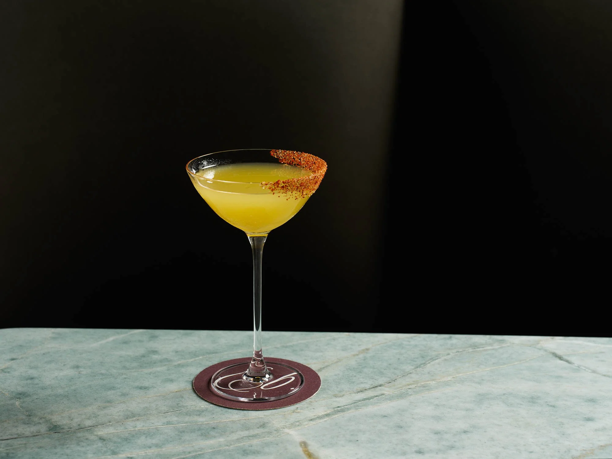 A yellow cocktail in a martini glass with a chili pepper rim, placed on a maroon coaster on a marble surface with a black background.