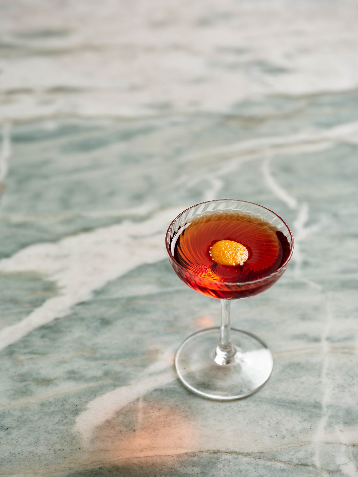 A glass of red cocktail with a yellow garnish, placed on a marble surface, with ocean waves in the background.