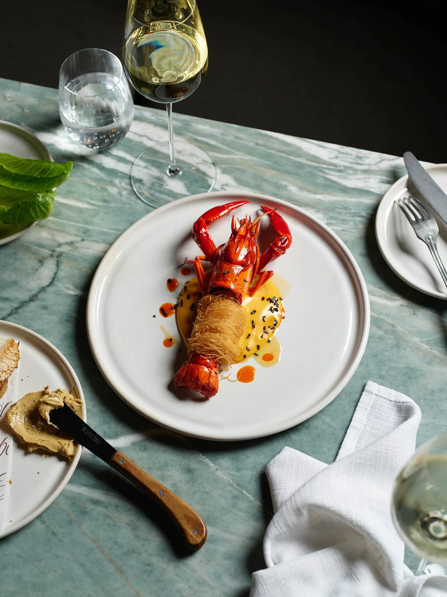 Lobster dish on a white plate with sauce and garnishes, surrounded by wine and water glasses on a marble table.