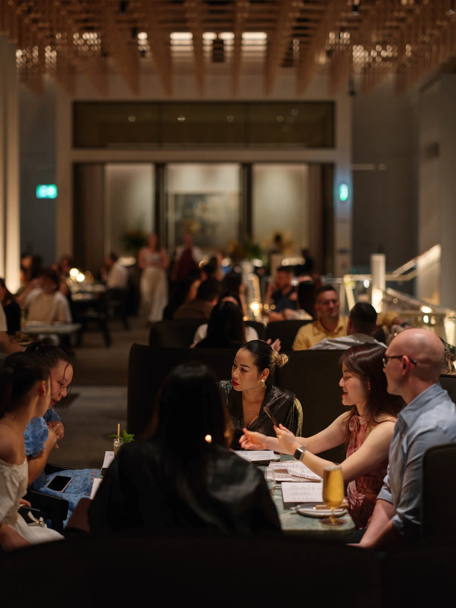 People dining at a restaurant with dim lighting, some looking at menus and engaging in conversation, with a busy background and elegant decor.