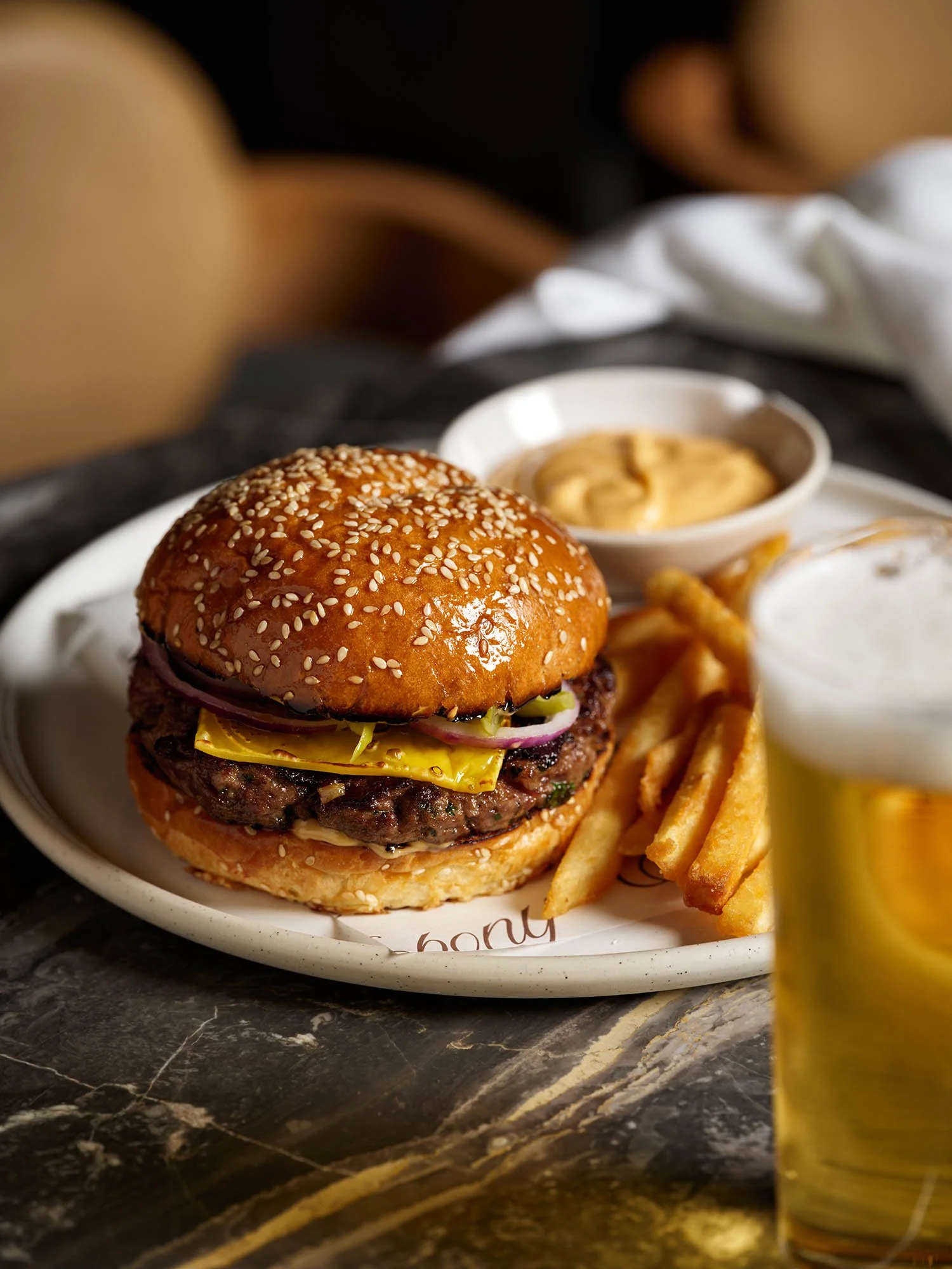 Close-up of a cheeseburger with sesame seed bun, lettuce, pickles, and onions, served with French fries, dipping sauces, and lemon on a white plate.