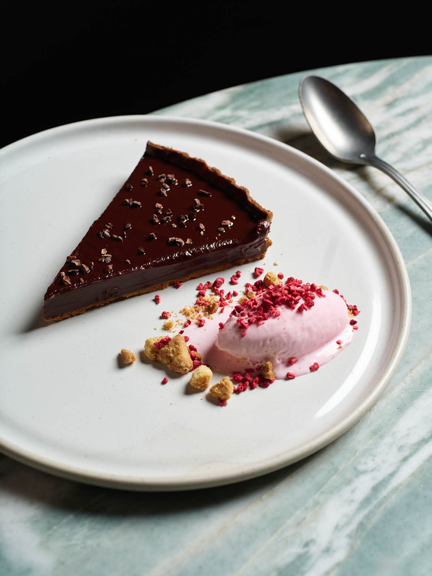 A white plate with a slice of chocolate tart and a dollop of pink ice cream topped with crushed red topping, garnished with cookie crumbs.