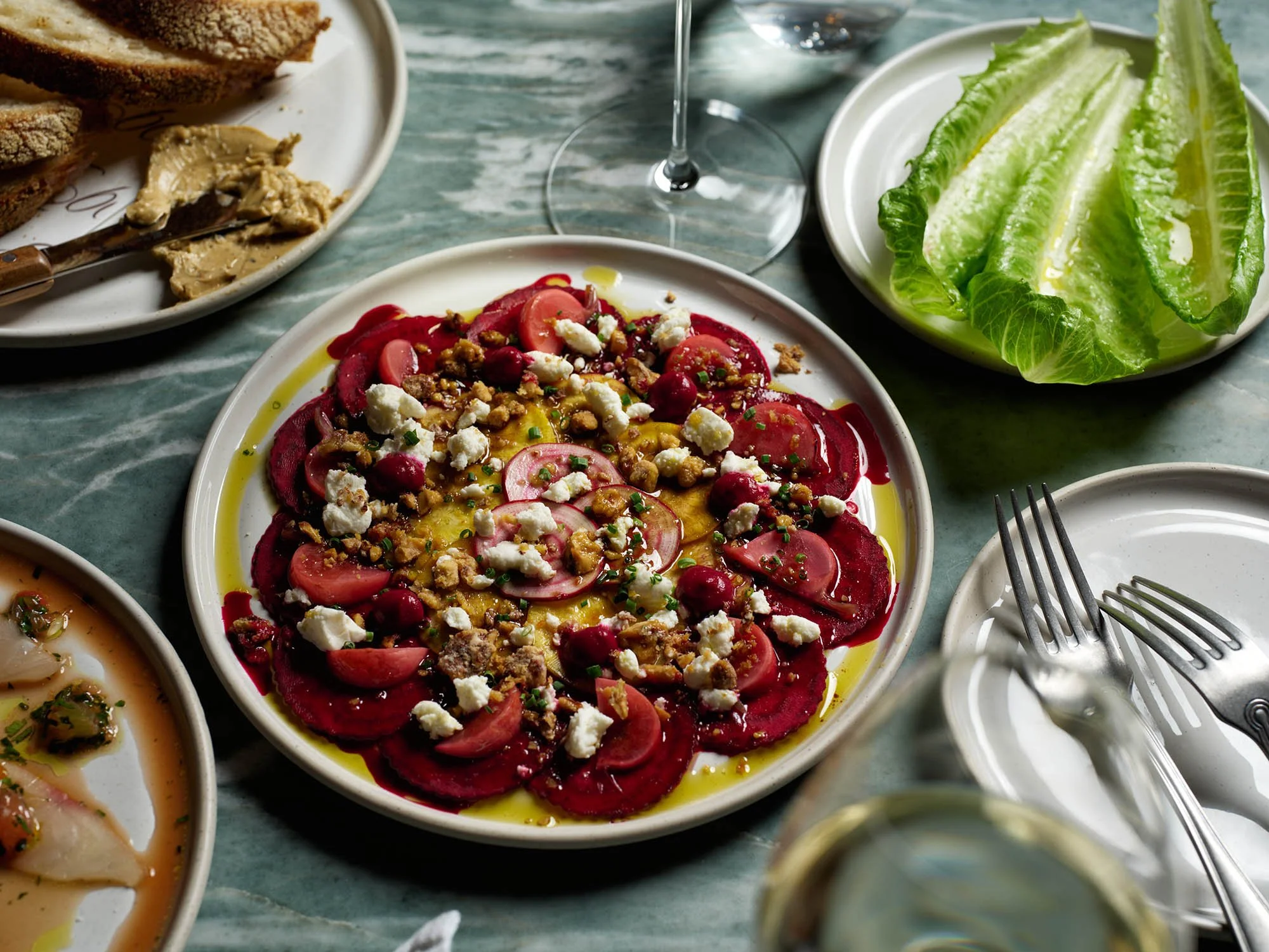 A plate of sliced radishes topped with crumbled cheese, chopped herbs, and a drizzle of oil, surrounded by other dishes and a glass of red wine on a marble table.