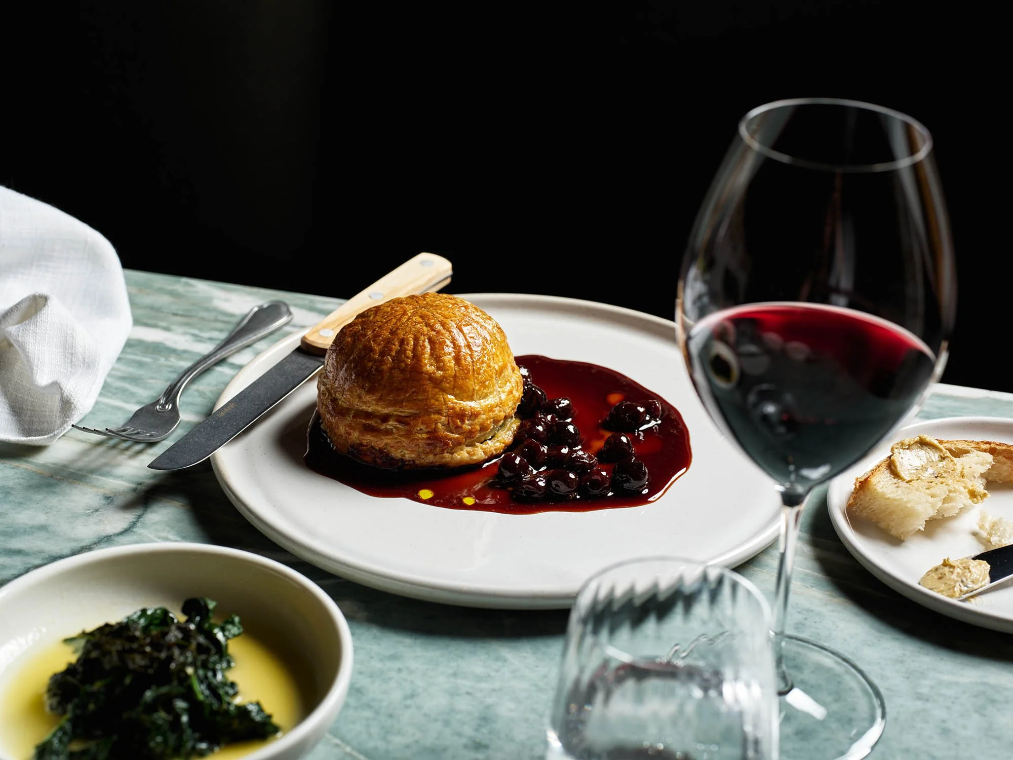 A plate with a baked pastry, berry compote, and a glass of red wine on a green marble table.