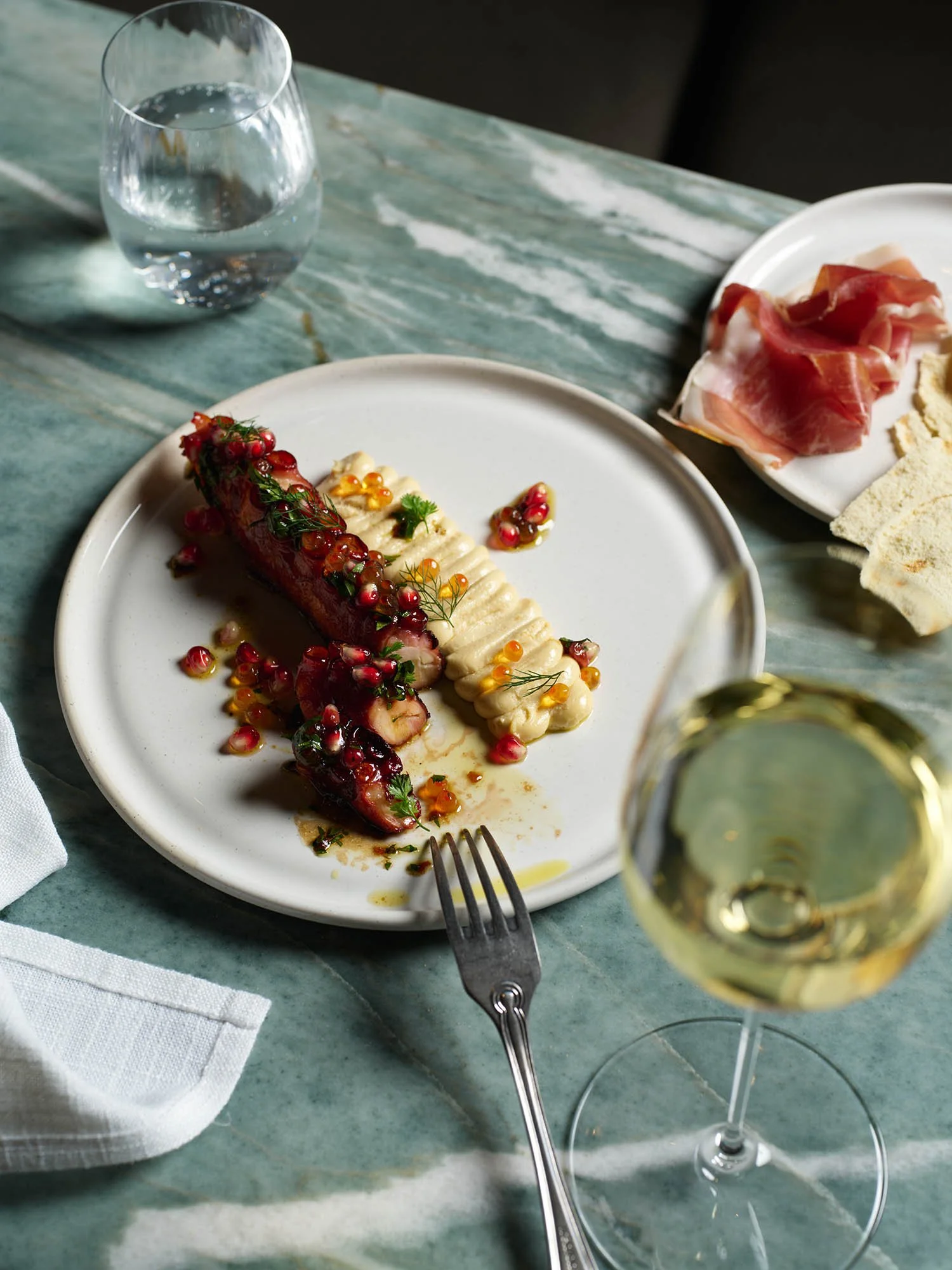Fremantle octopus with whipped cod roe, pomegranate vinaigrette, paired with a glass of white wine, on a marble table with water and charcuterie in the background.