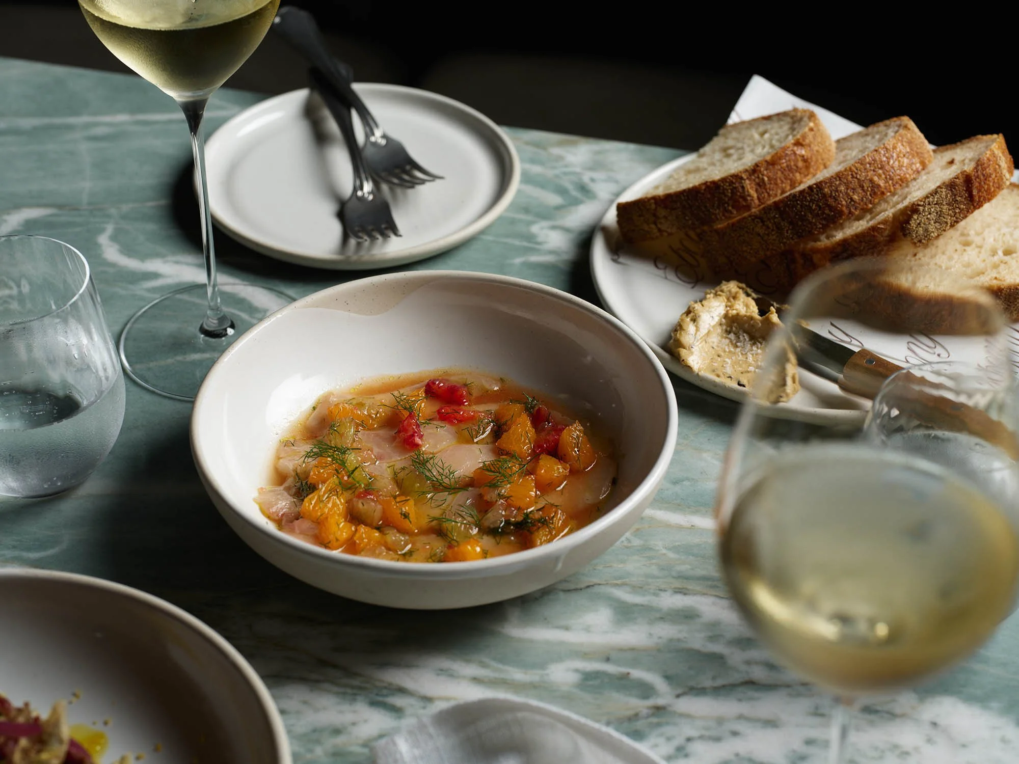 Table setting with a bowl of fish with garnishes, a glass of white wine, a glass of water, and plates of sliced bread with a spread, on a marble table.
