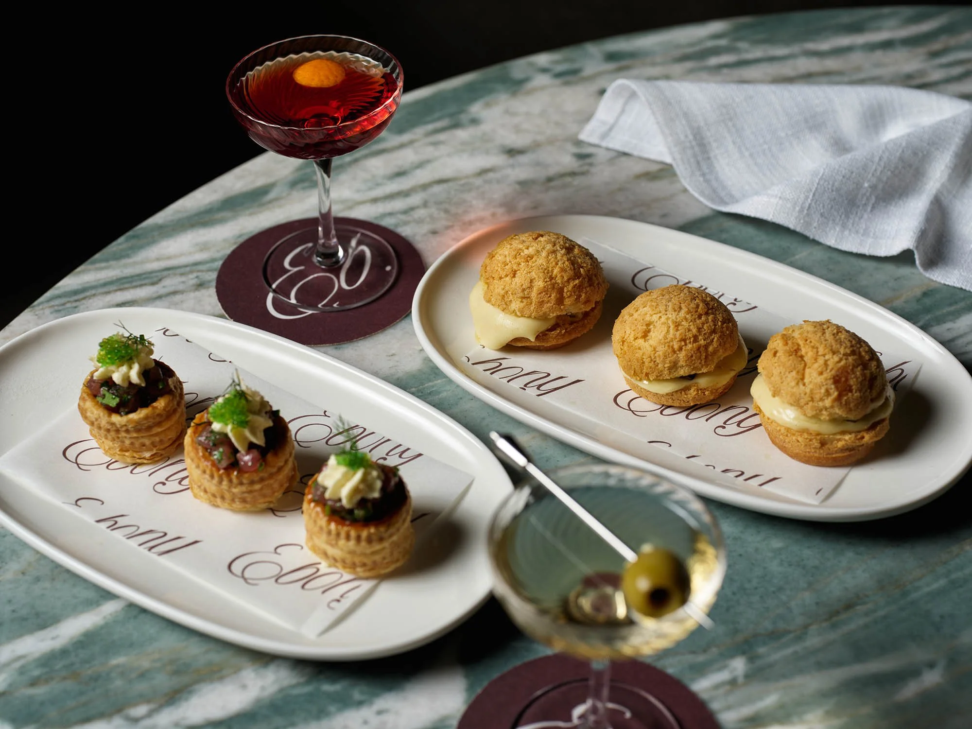 Assorted appetizers and cocktails on a marble table, including three small savory pastries with toppings, three cream-filled choux buns, a glass of red wine, and a martini with an olive garnish.