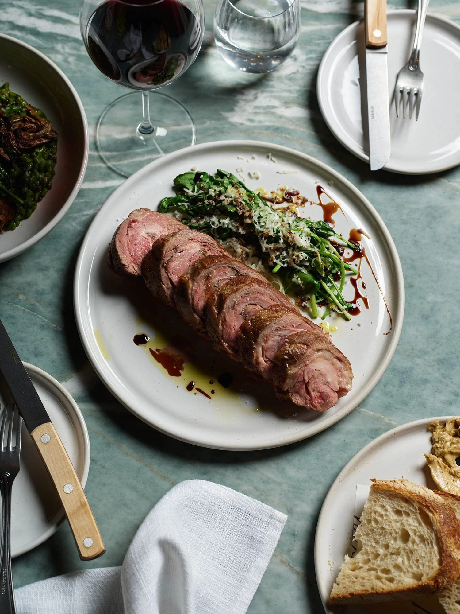 A plated meal featuring sliced beef tenderloin with a side salad of greens, grated cheese, and balsamic drizzle, along with a glass of red wine, a glass of water, and some bread on a separate plate, set on a marble table.
