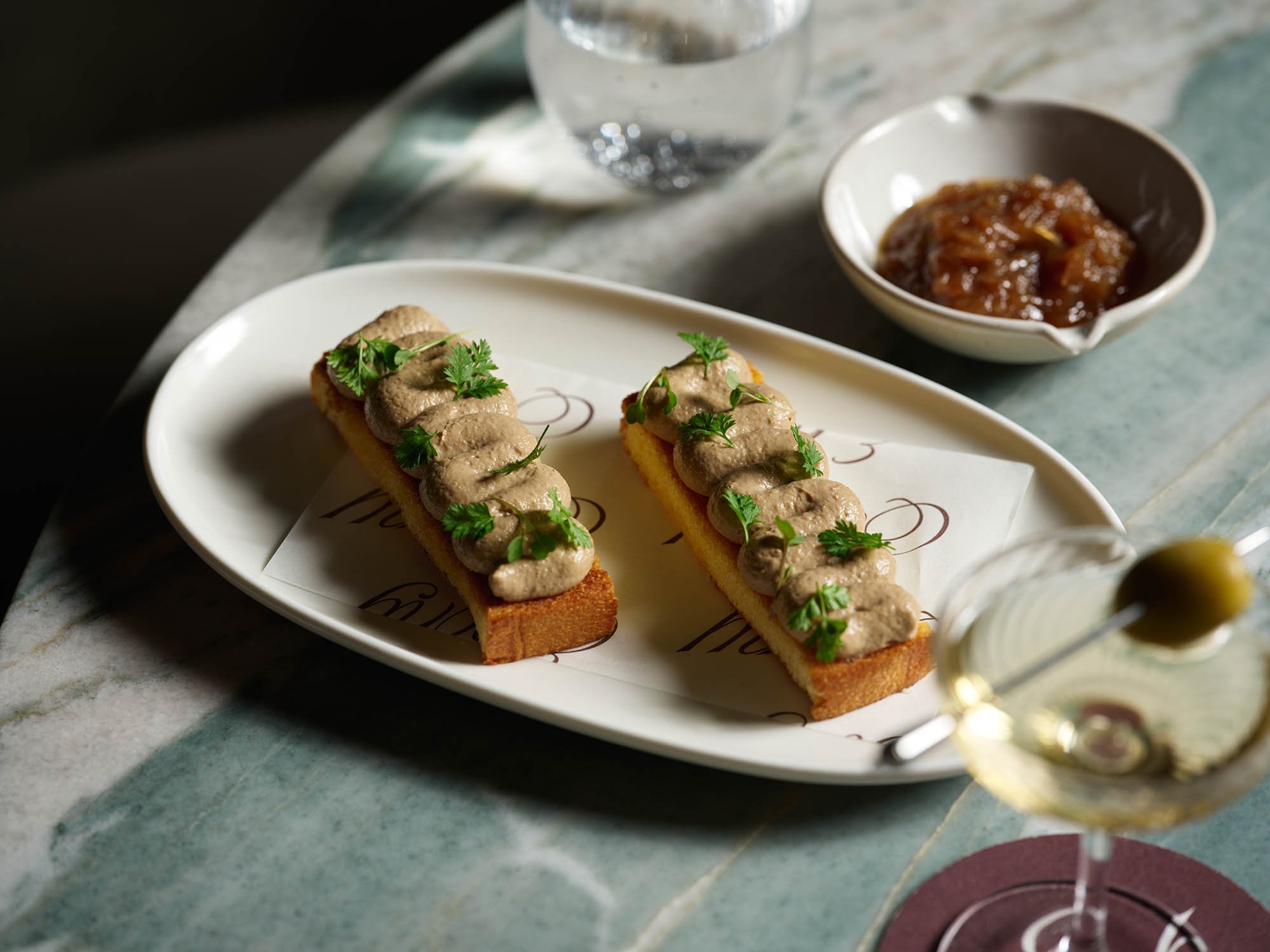 Two pieces of toast topped with mushroom or liver pâté garnished with parsley, served on a white oval plate with a printed paper liner, accompanied by a glass of water, a small bowl of caramelized onions or chutney, and a glass of white wine on a mar