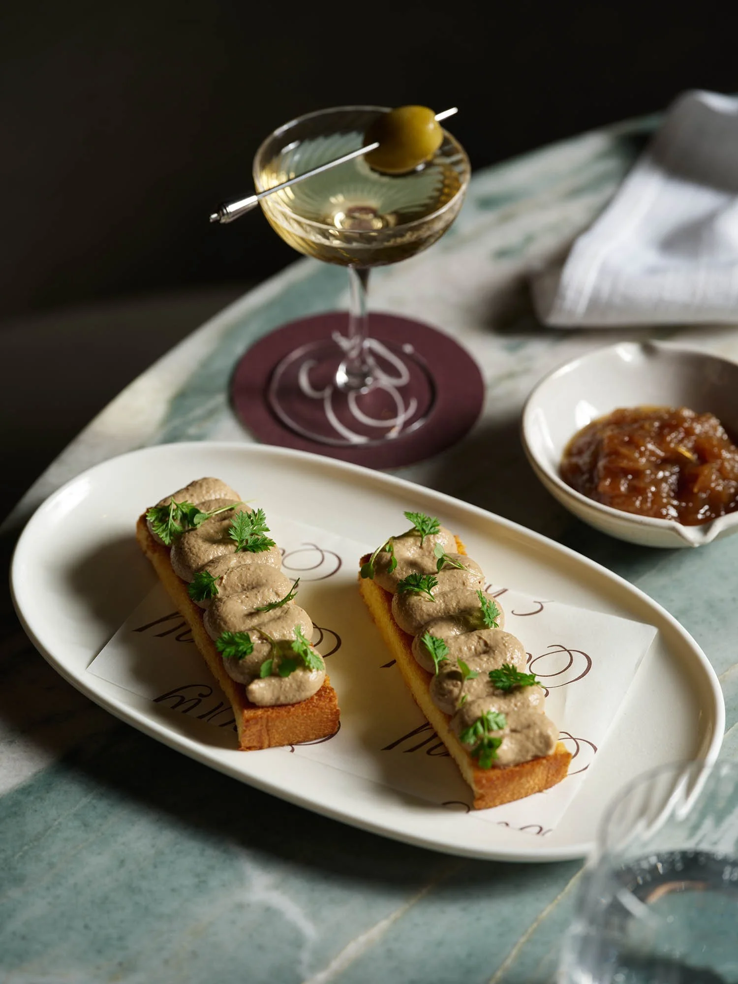 Two slices of baguette topped with pâté and garnished with fresh herbs on a white plate, a glass of cocktail with an olive, a bowl of chutney or relish, and a folded napkin on a marble table.