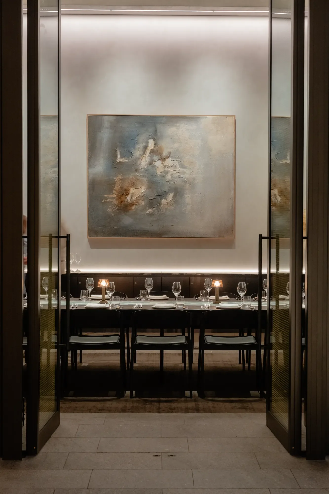 Elegant dining area with a table set for a meal, featuring wine glasses, a small table lamp, and modern artwork on the wall, viewed through an open pair of glass doors.