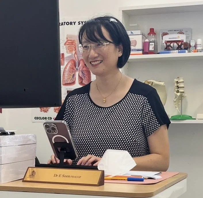 A cheerful woman with short black hair and glasses, sitting at a desk in a medical office, wearing a black and white polka dot shirt, with a nameplate that reads Dr Elaine Soon, surrounded by medical supplies and anatomical models.