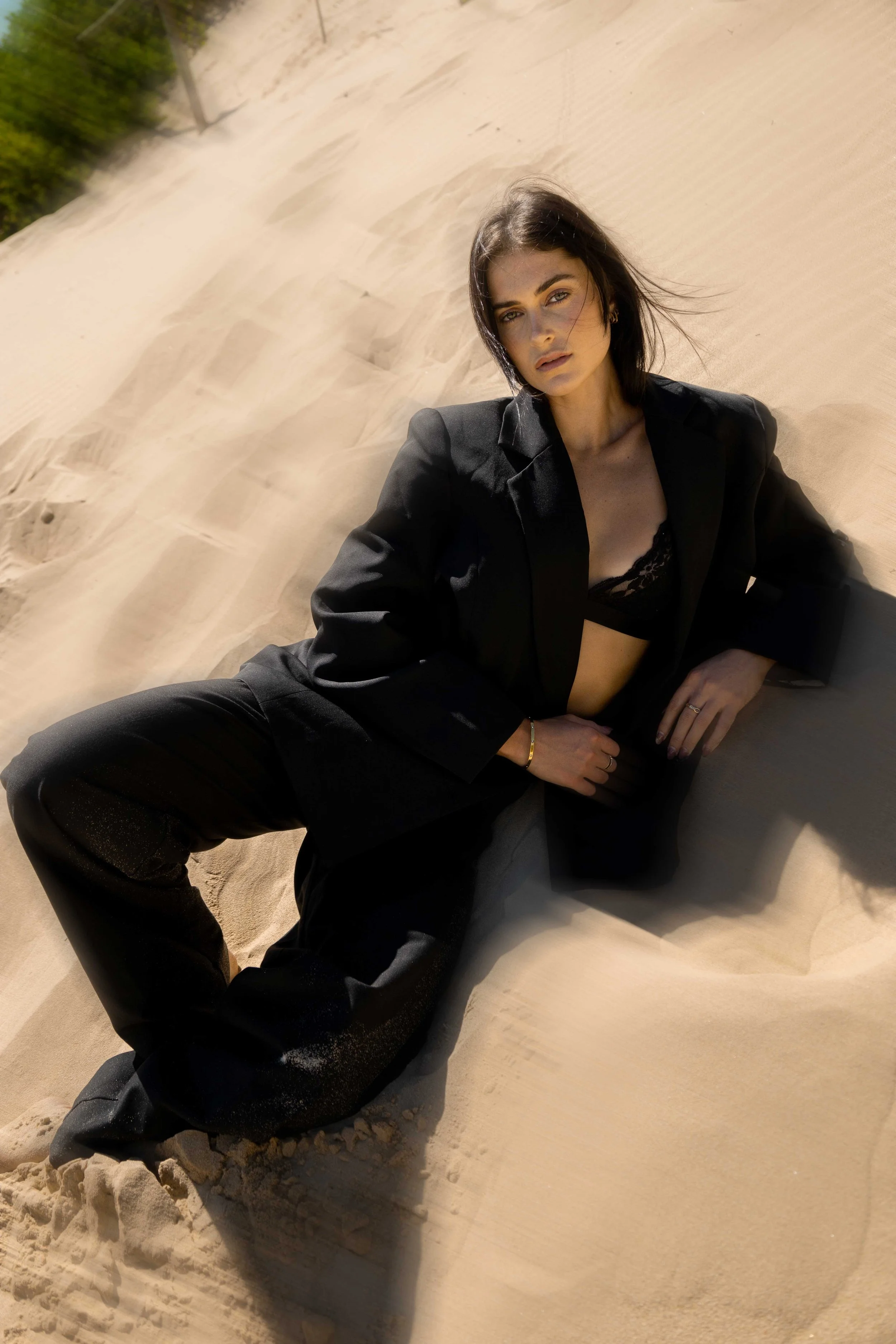 A woman dressed in a black blazer and lace top, sitting on sand dunes with sunlight shining on her face, in a desert setting.