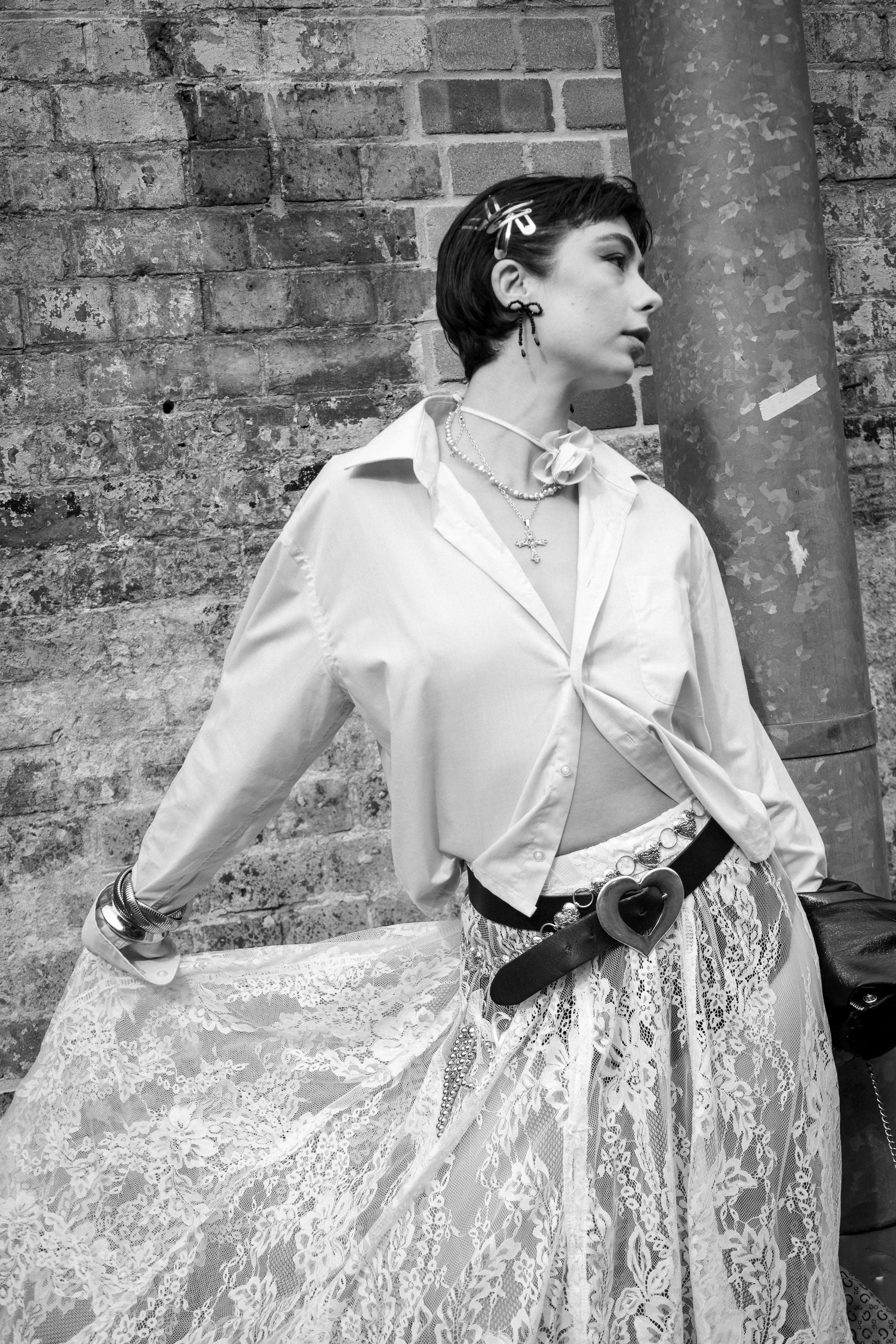 A young woman with short dark hair styled with clips, wearing earrings, necklaces, a button-up shirt tied at the waist, and a lace skirt with a wide belt featuring a heart-shaped buckle, leaning against a brick wall and a metal pipe.