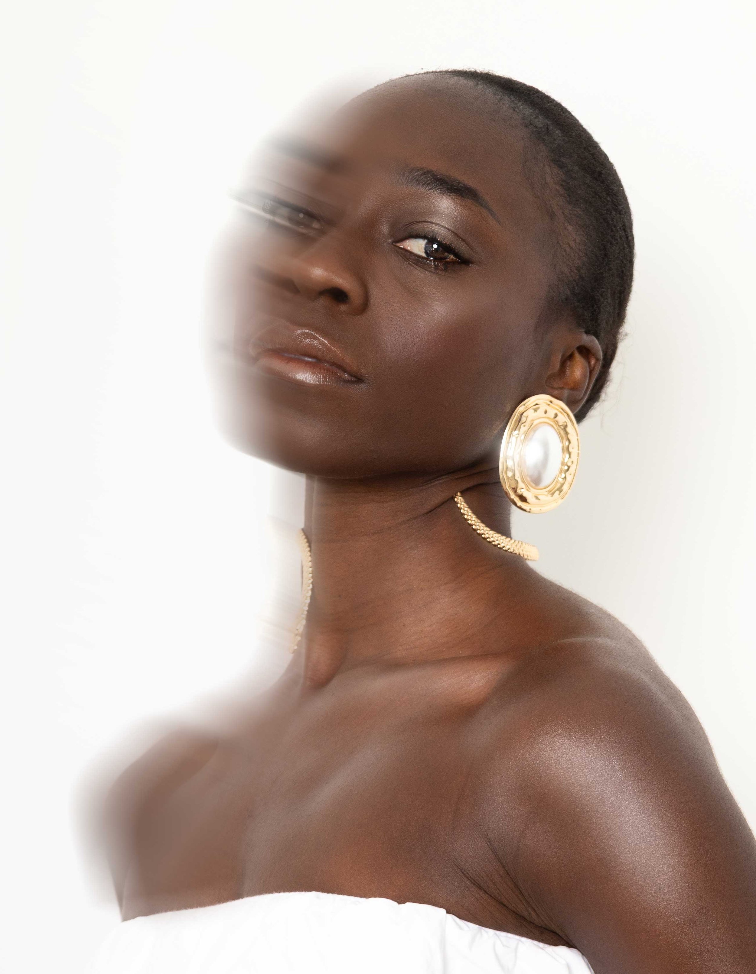 Close-up portrait of a woman with dark skin, short hair, and wearing large gold and pearl earrings. She has subtle makeup and is dressed in a white off-the-shoulder top. The background is plain white.