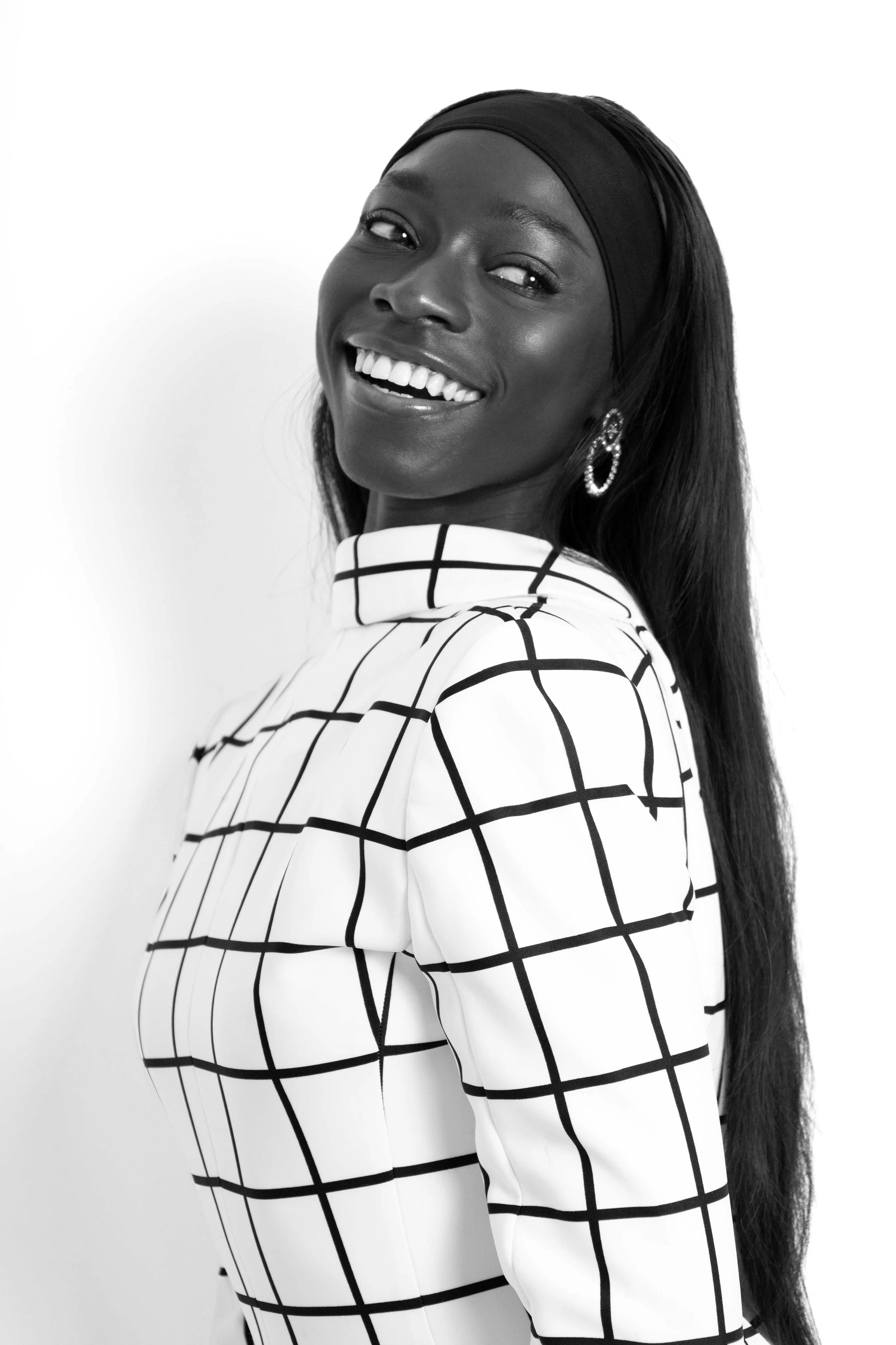 Black and white photograph of a smiling Black woman with long hair, wearing a headband and a checkered top, posing against a plain background.