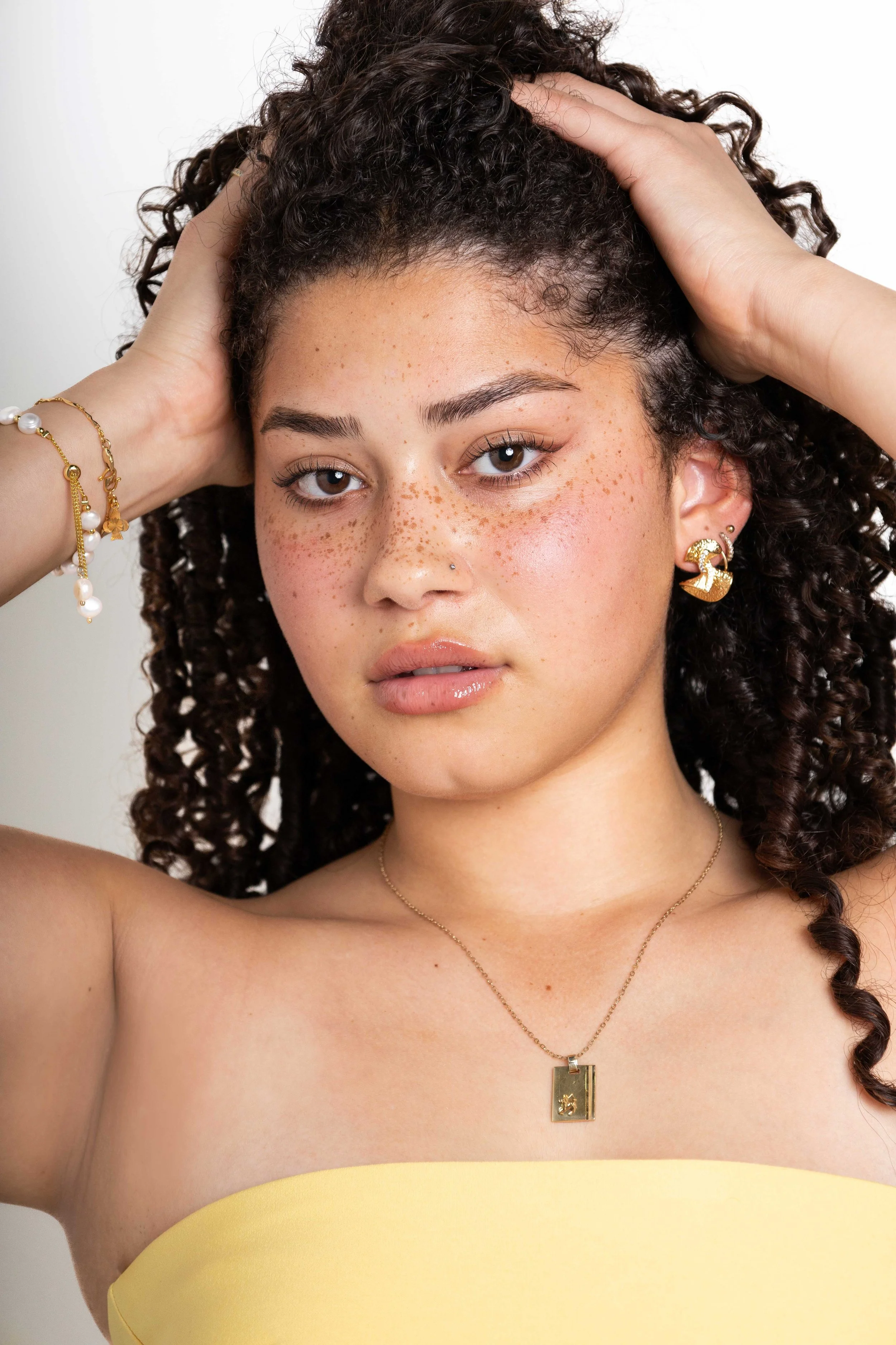 A young woman with curly hair and light skin, wearing gold jewelry, including earrings, a necklace, and bracelets, posing with her hands on her head and looking at the camera.