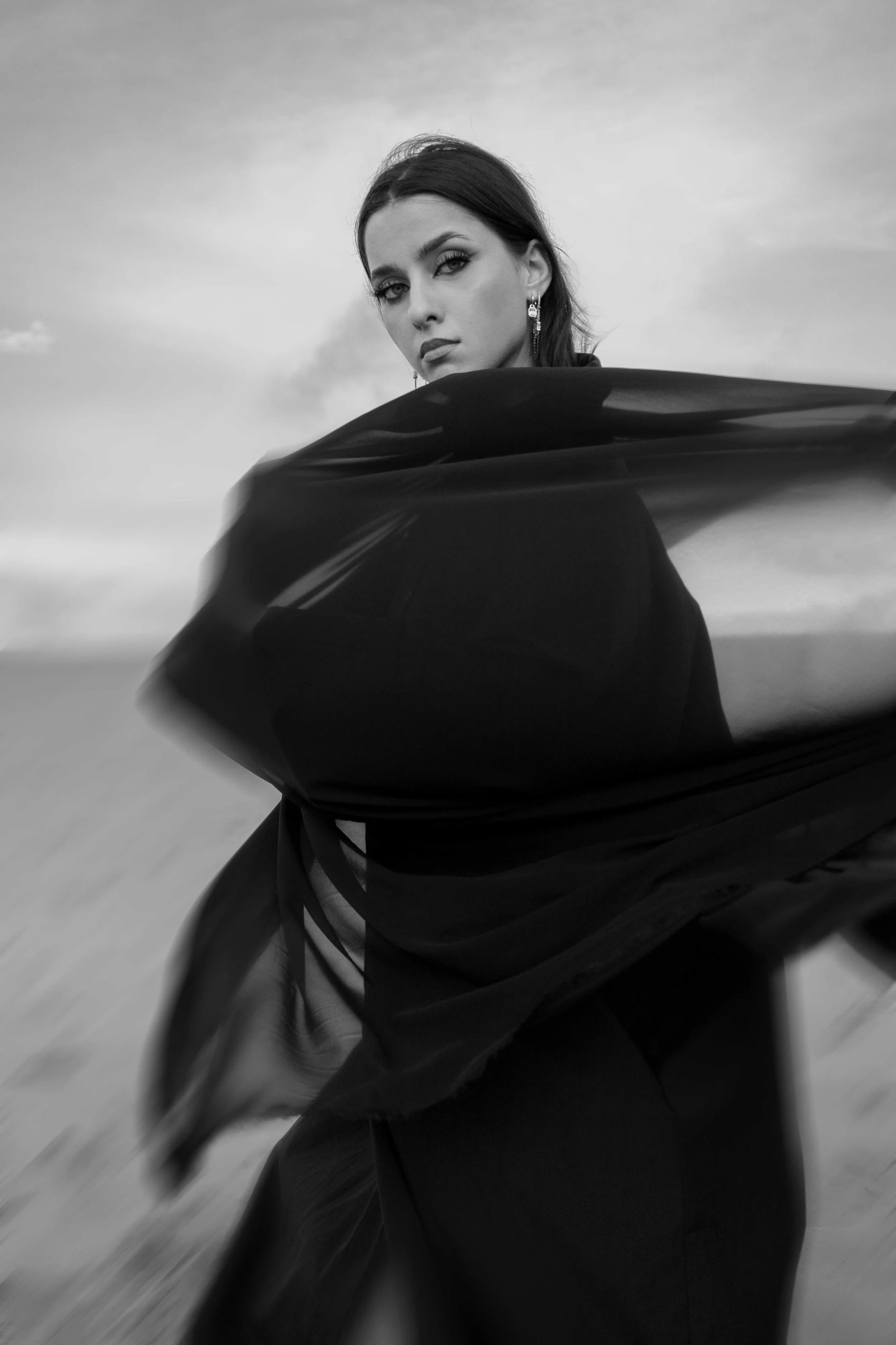 A black and white photo of a woman with dark hair and earrings, looking at the camera, with a flowing black garment in motion.