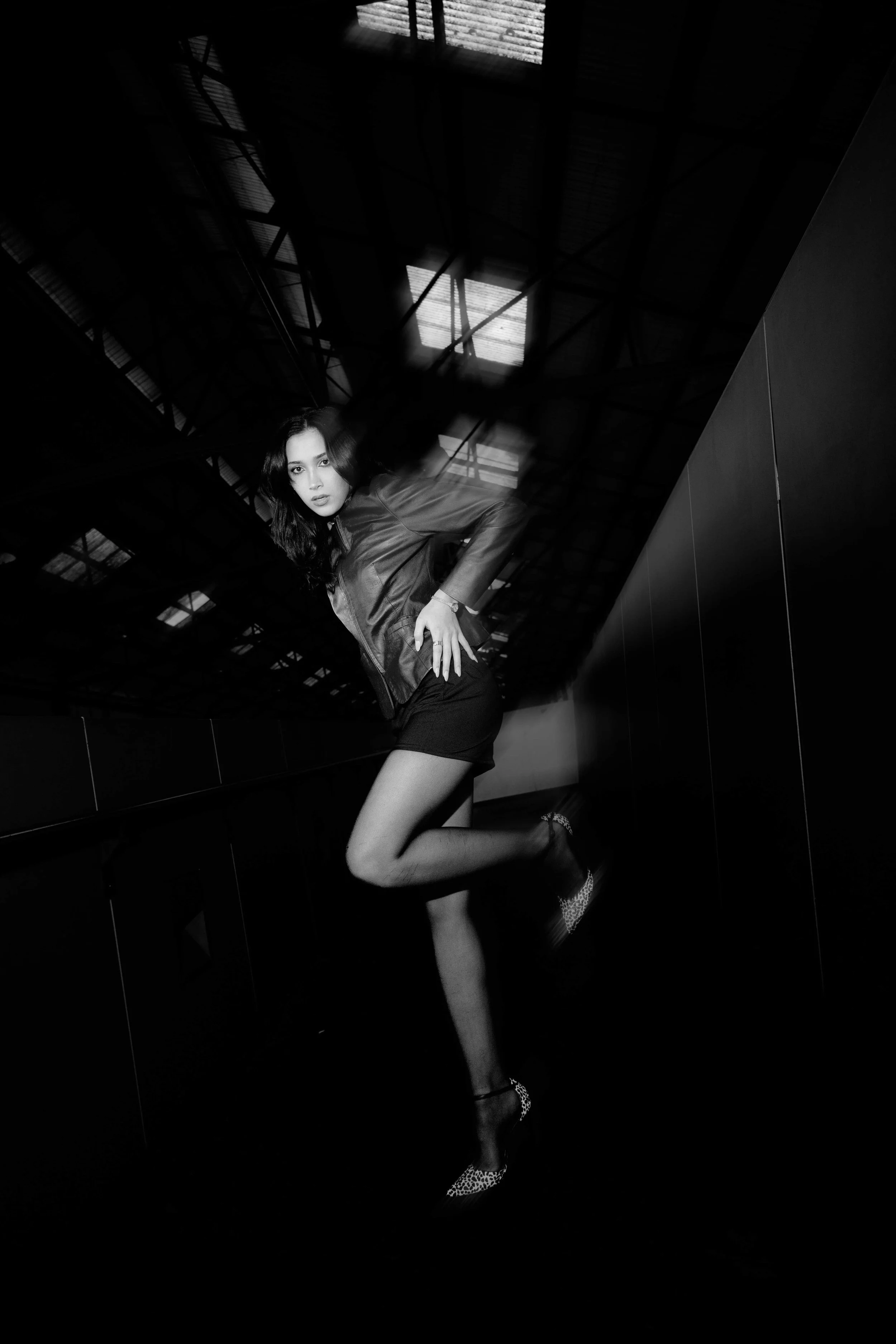 Black and white photo of a woman in a leather jacket, short skirt, and leopard print heels, posing indoors under a high ceiling with bright light coming through windows.