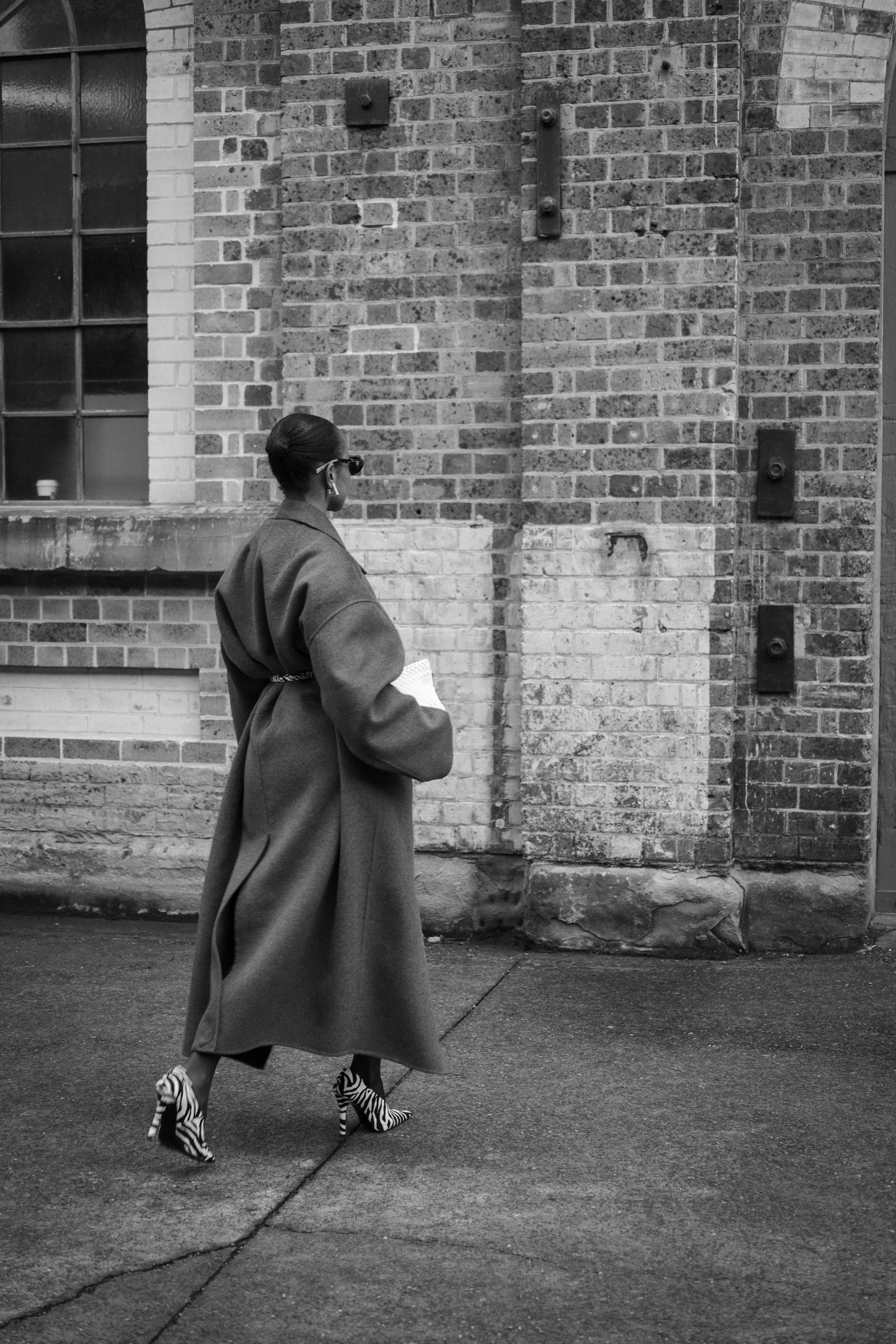 A woman walking along a sidewalk near a brick wall, dressed in a long coat, zebra-striped high heels, and sunglasses.