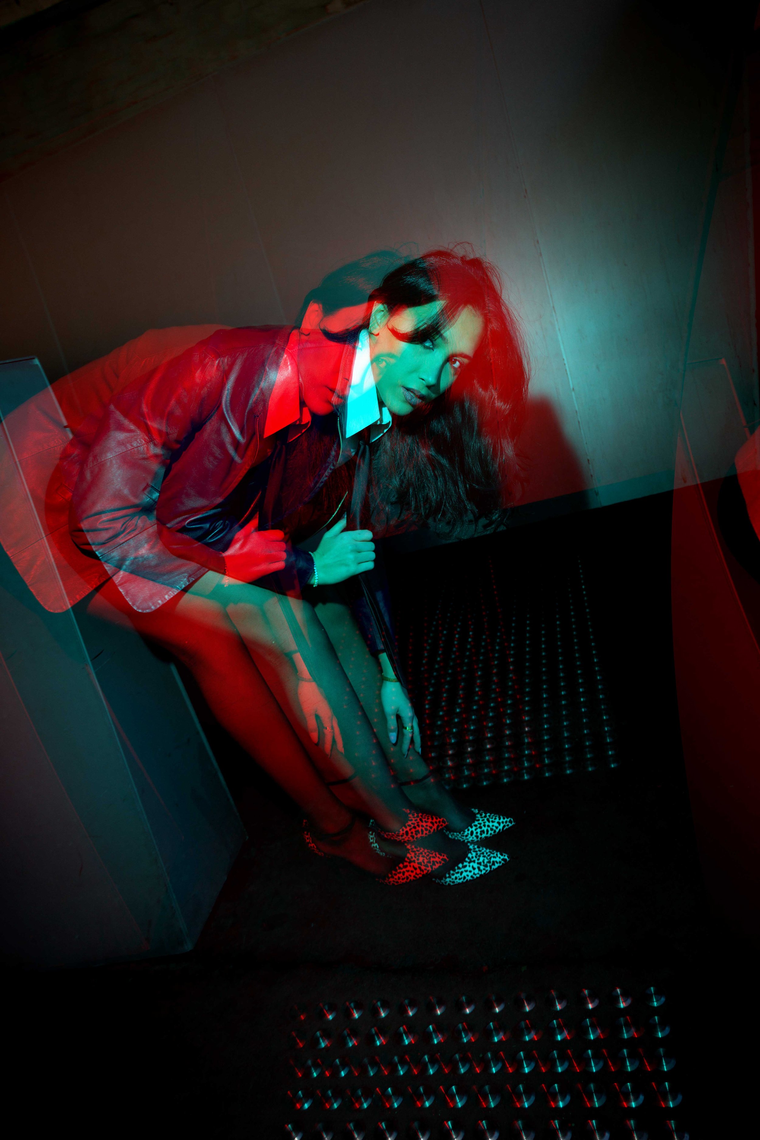 A woman with long dark hair, wearing a leather jacket, blouse, and patterned high heels, sitting on a bench in a dimly lit room with colorful lighting and a textured black floor.