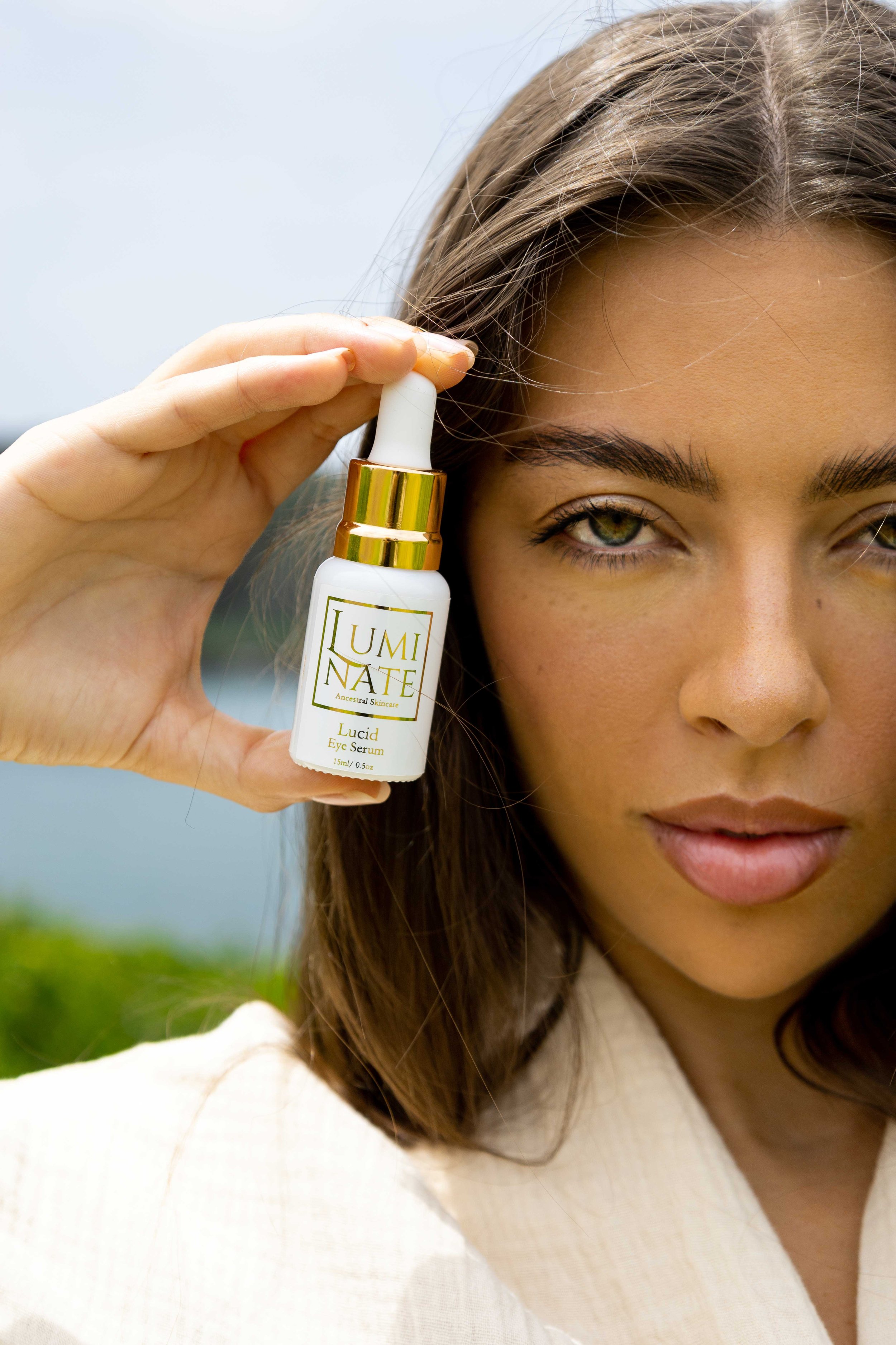 A woman with long brown hair and green eyes holds a small bottle of 'Luminate Lucid Eye Serum' close to her face, outdoors during daytime.